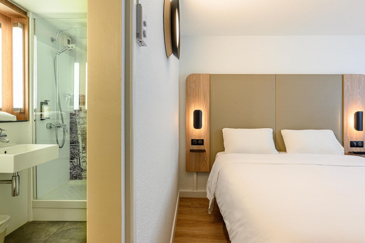 Habitación de hotel con ducha, lavabo, cama y lámpara fijada a la pared.