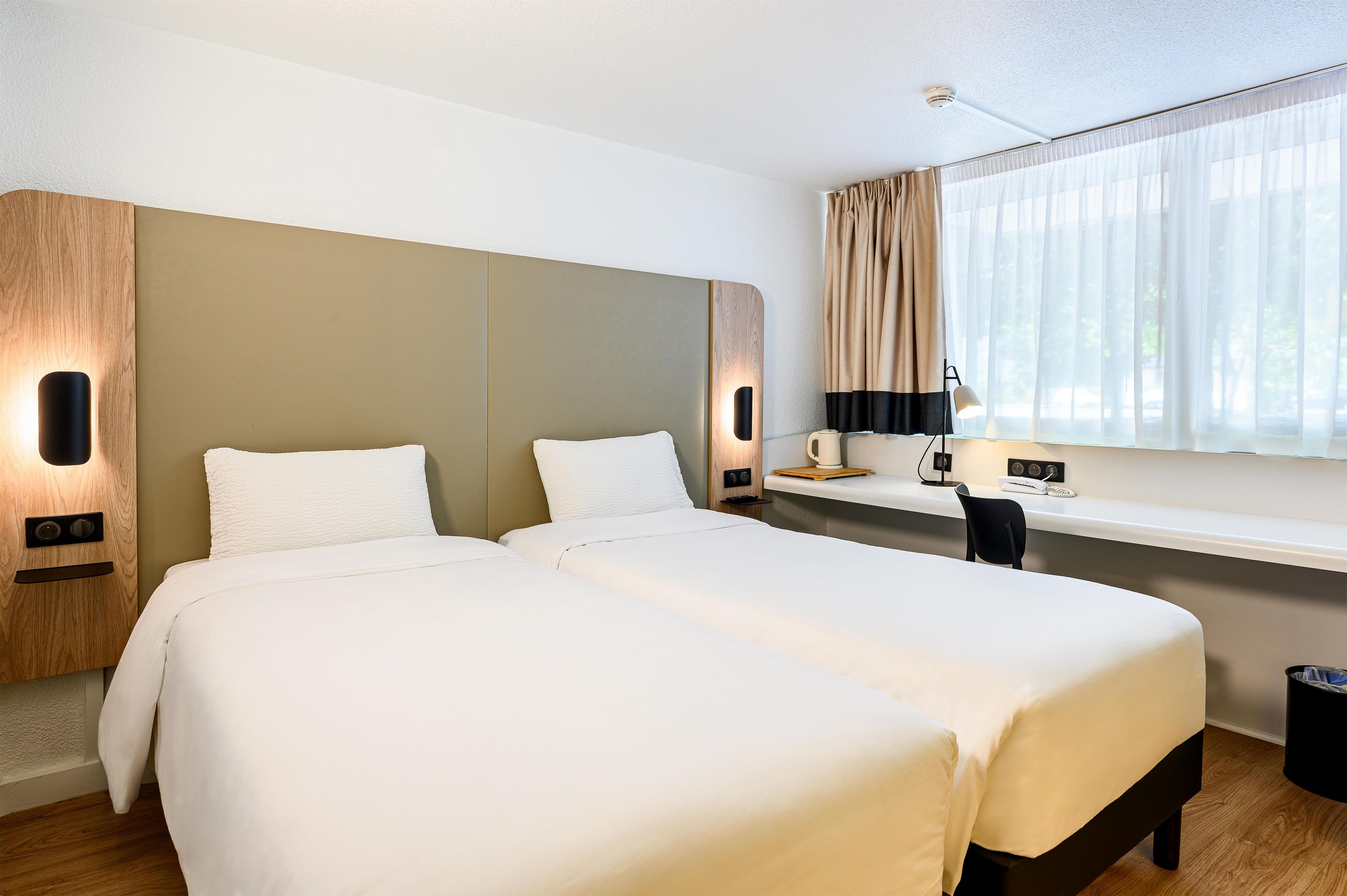 A hotel room with two beds, a desk, a chair, a lamp, and a window with curtains.