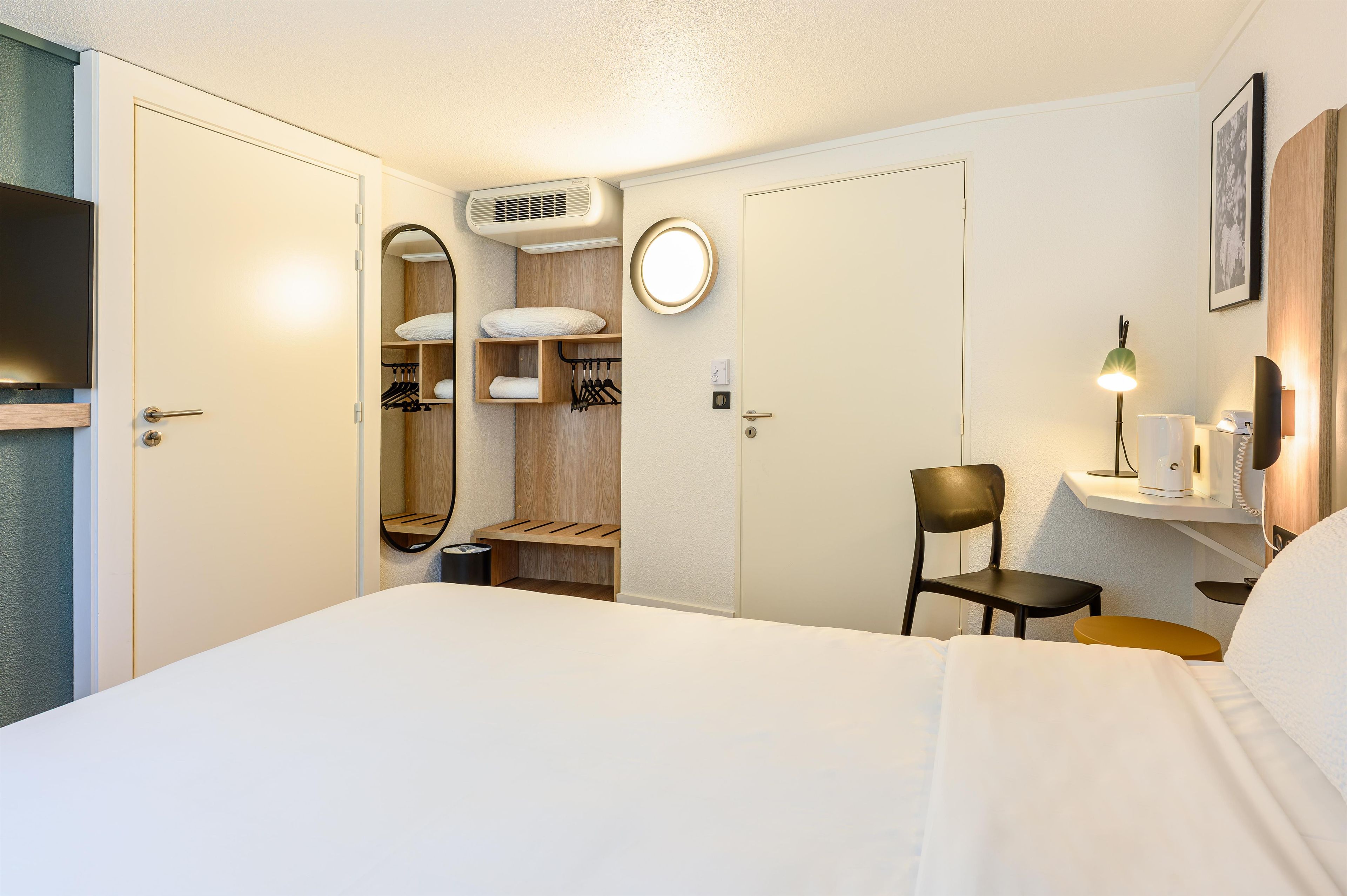 Een hotelkamer met een bed, een stoel, een bureau met een lamp, een spiegel, een kast en een klein plankje met een telefoon.