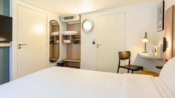 Een hotelkamer met een bed, een stoel, een bureau met een lamp, een spiegel, een kast en een klein plankje met een telefoon.