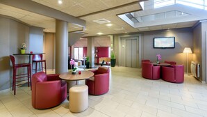 Un hall d’entrée spacieux avec un plafond haut, un espace salon central agrémenté de fauteuils et de poufs rouges, une petite table ornée d’un vase de fleurs et un téléviseur fixé au mur.