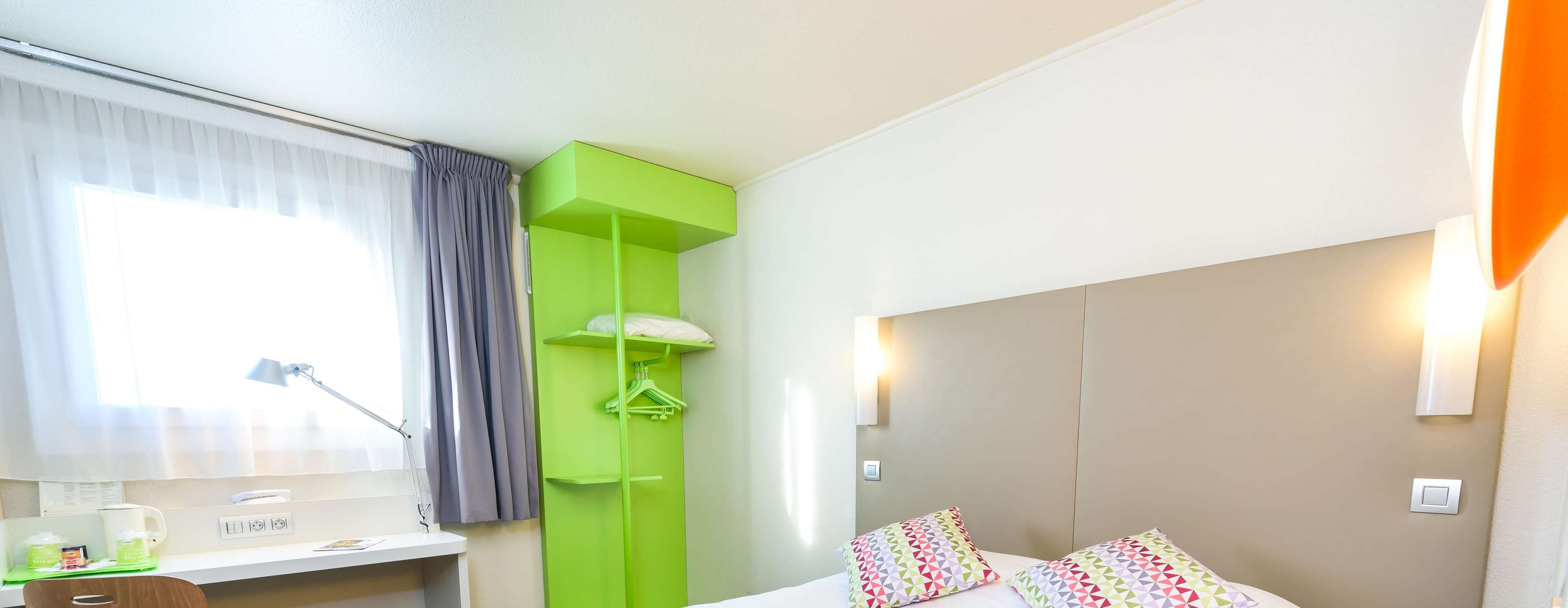 Ett hotellrum med en säng, ett skrivbord med en lampa, en grön garderob och ett fönster med gardiner.