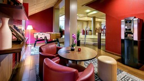 Un hall d’hôtel avec une table ronde, des chaises rouges, un canapé, un réfrigérateur et une bibliothèque.