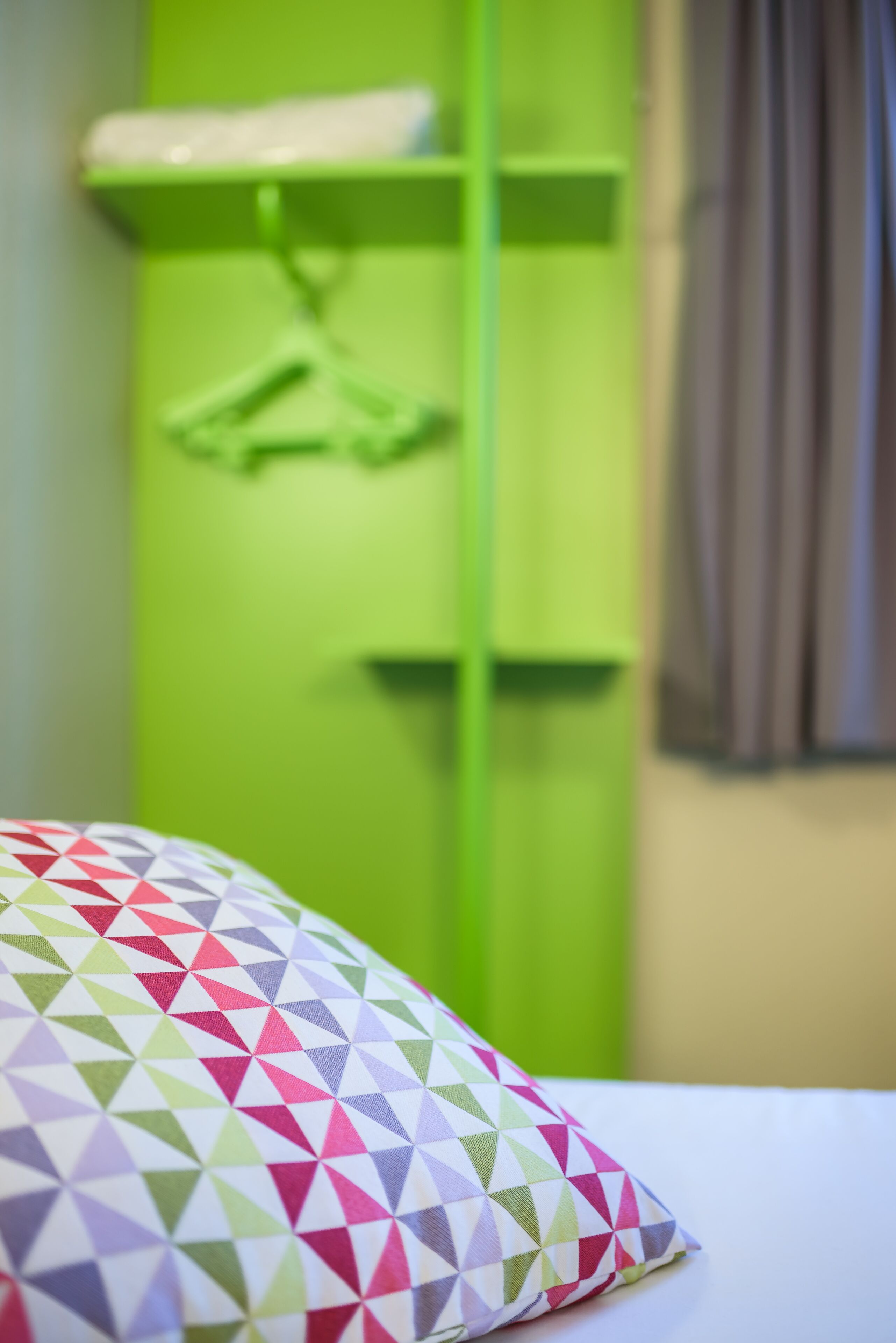 A bed with a patterned pillow, a green wardrobe, and a window with curtains.