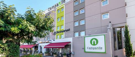 A hotel with a green and gray facade, outdoor seating area, and a sign indicating it is a hotel and restaurant.