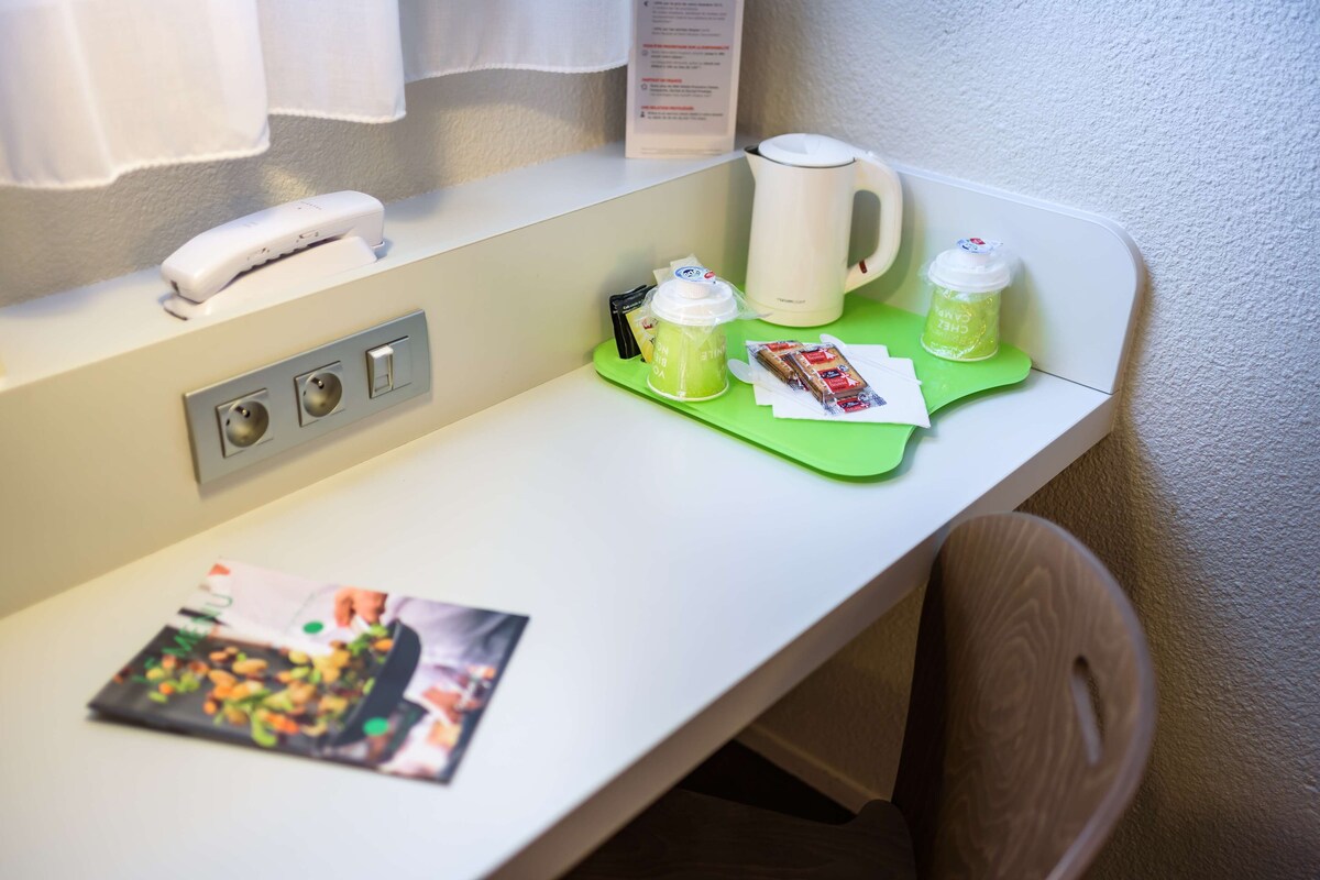 Ein Hotelzimmer-Tresen mit einer weißen Kaffeemaschine, einem weißen Telefon, einer grünen Ablage mit Tasse und kleiner Verpackung sowie einer Zeitschrift auf dem Tresen.