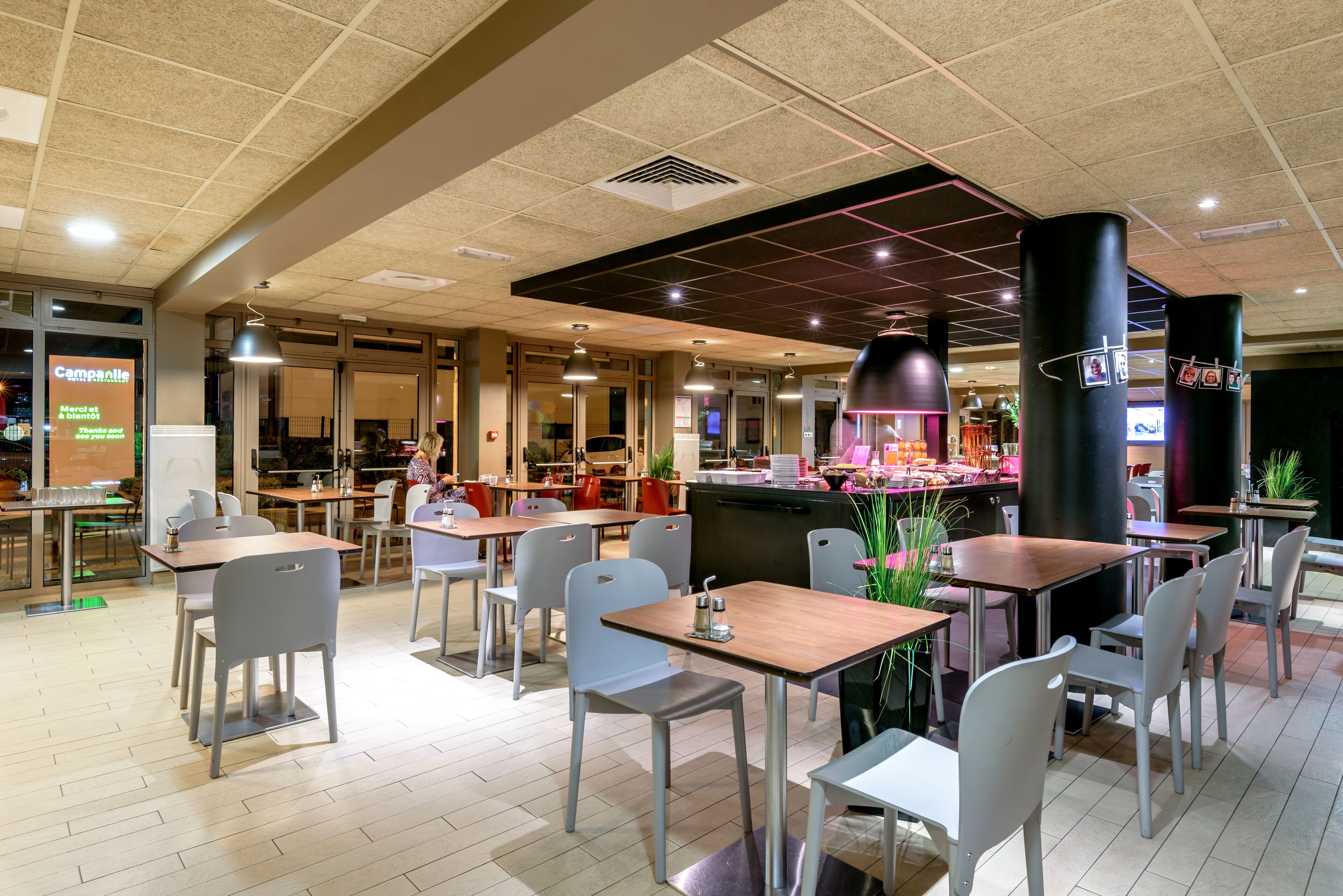 A modern restaurant with multiple dining tables and chairs, a counter with a menu display, and a ceiling with a grid pattern.