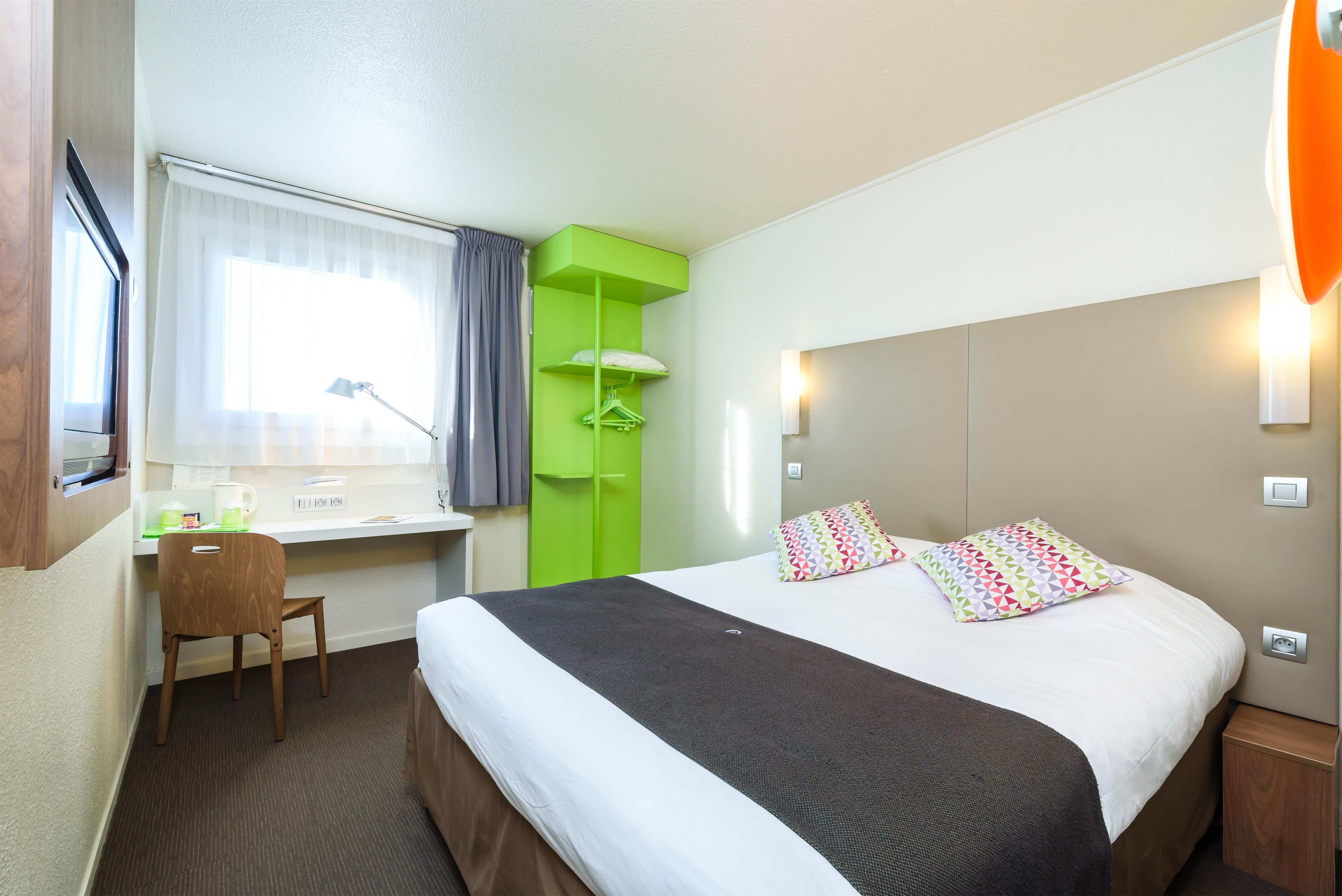 A hotel room with a bed, a desk, a chair, a window with curtains, and a green wardrobe.