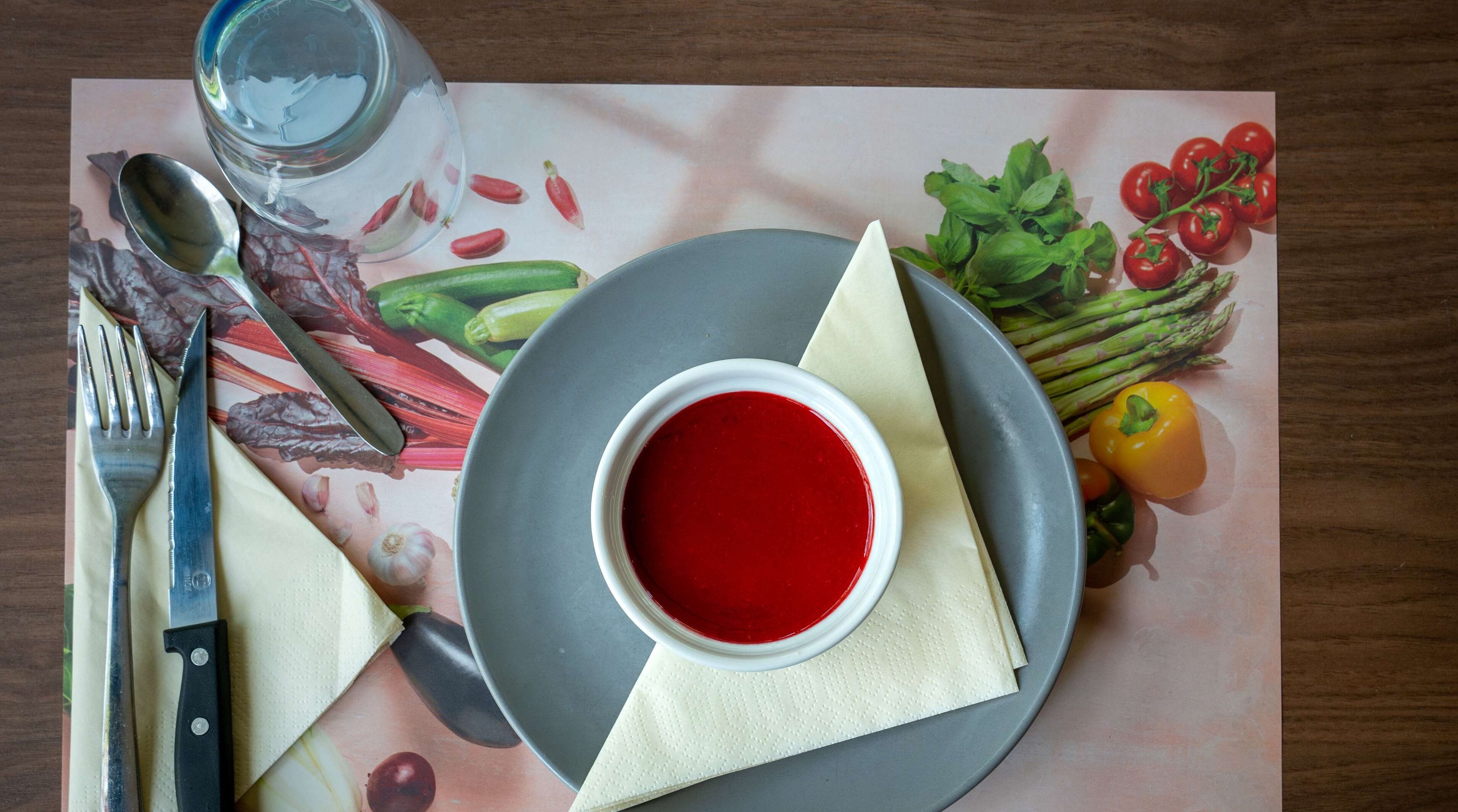 A table setting with a bowl of red soup, a glass of water, a knife, a fork, a spoon, and various vegetables.