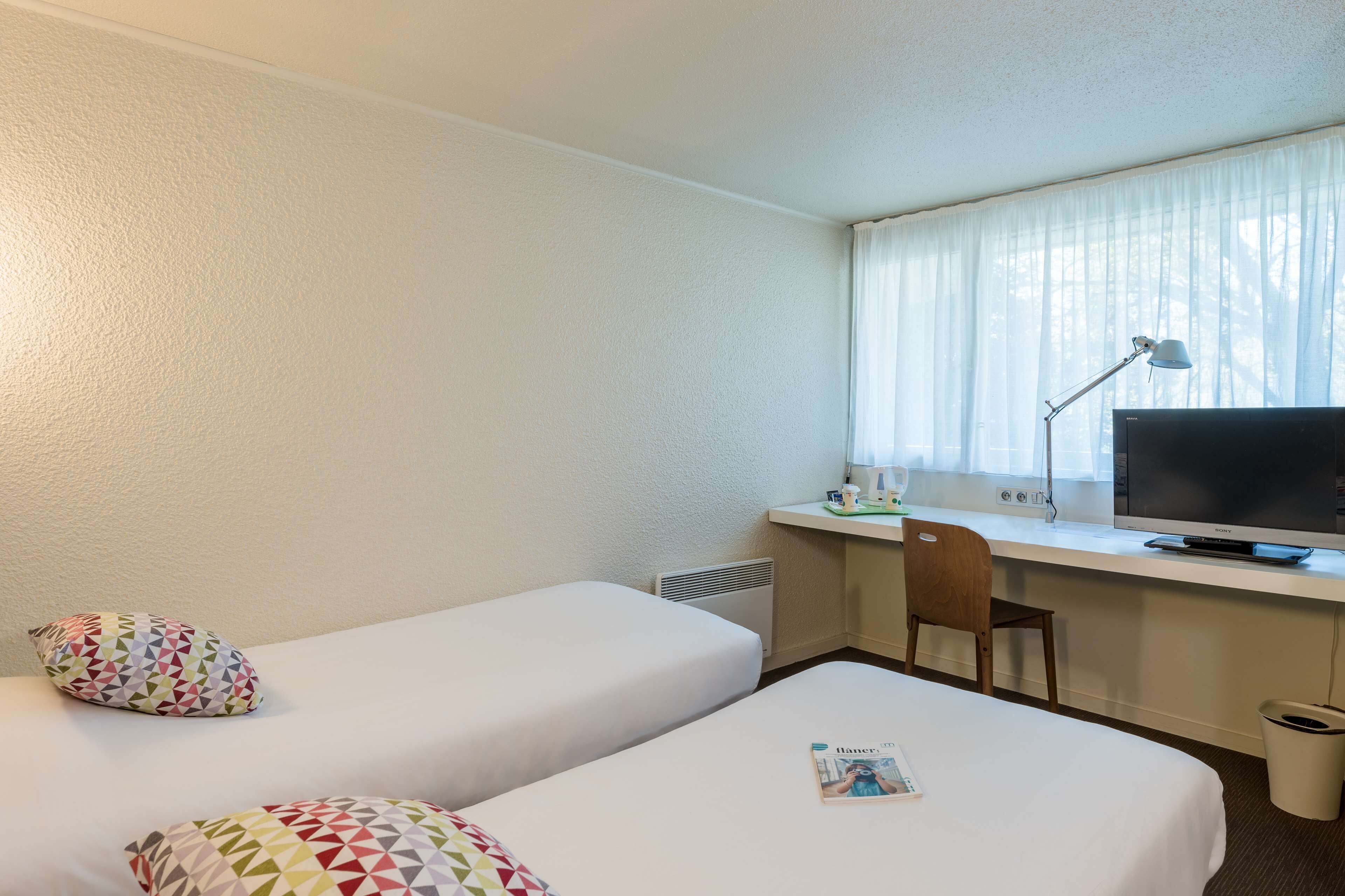 A hotel room with two beds, a desk with a computer and a lamp, a chair, and a window with curtains.