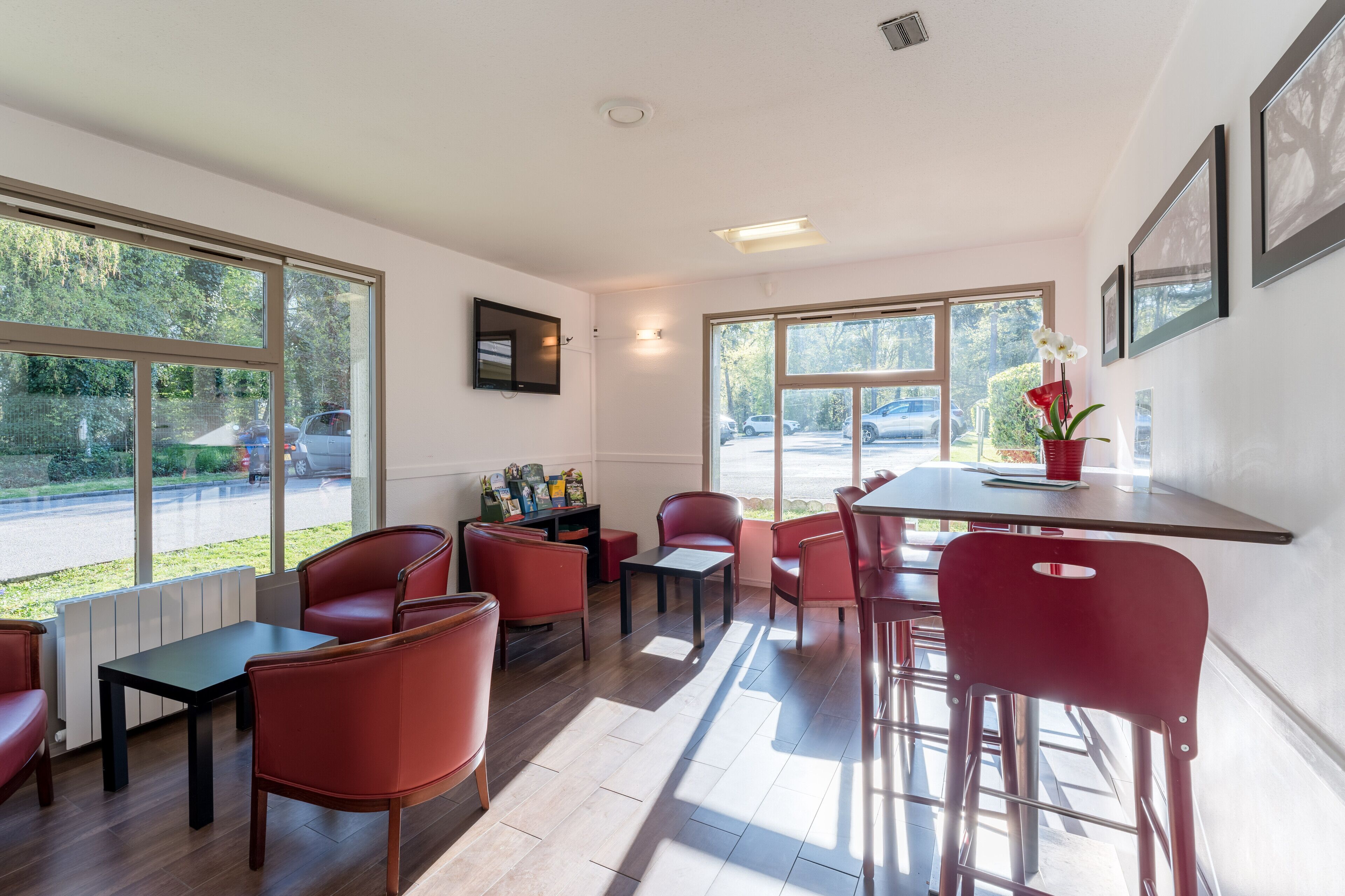 A modern dining area with red chairs and tables, large windows with a view of the parking lot, and a wall-mounted TV.