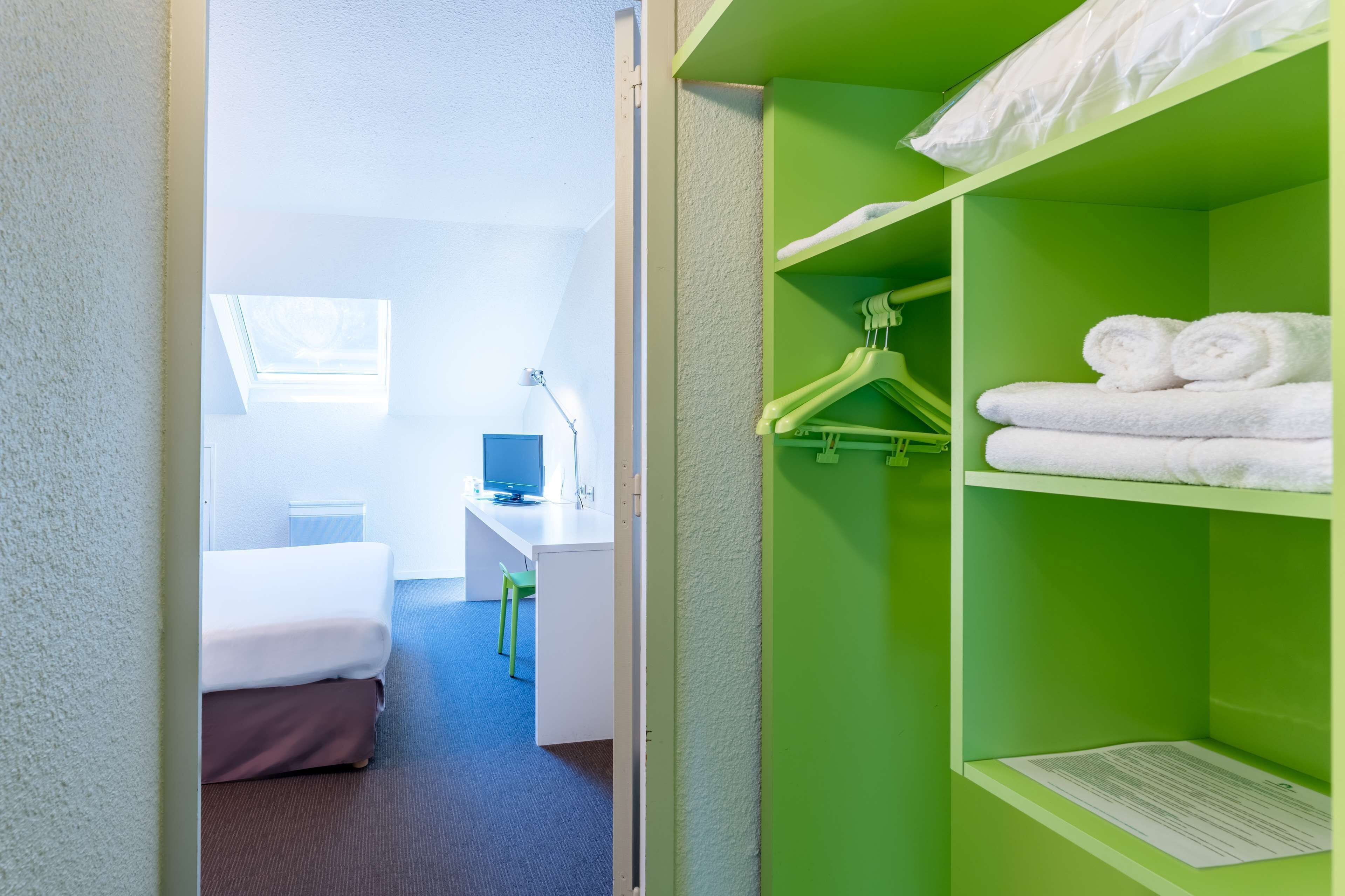 A hotel room with a bed, desk, and green wardrobe with hanging clothes.