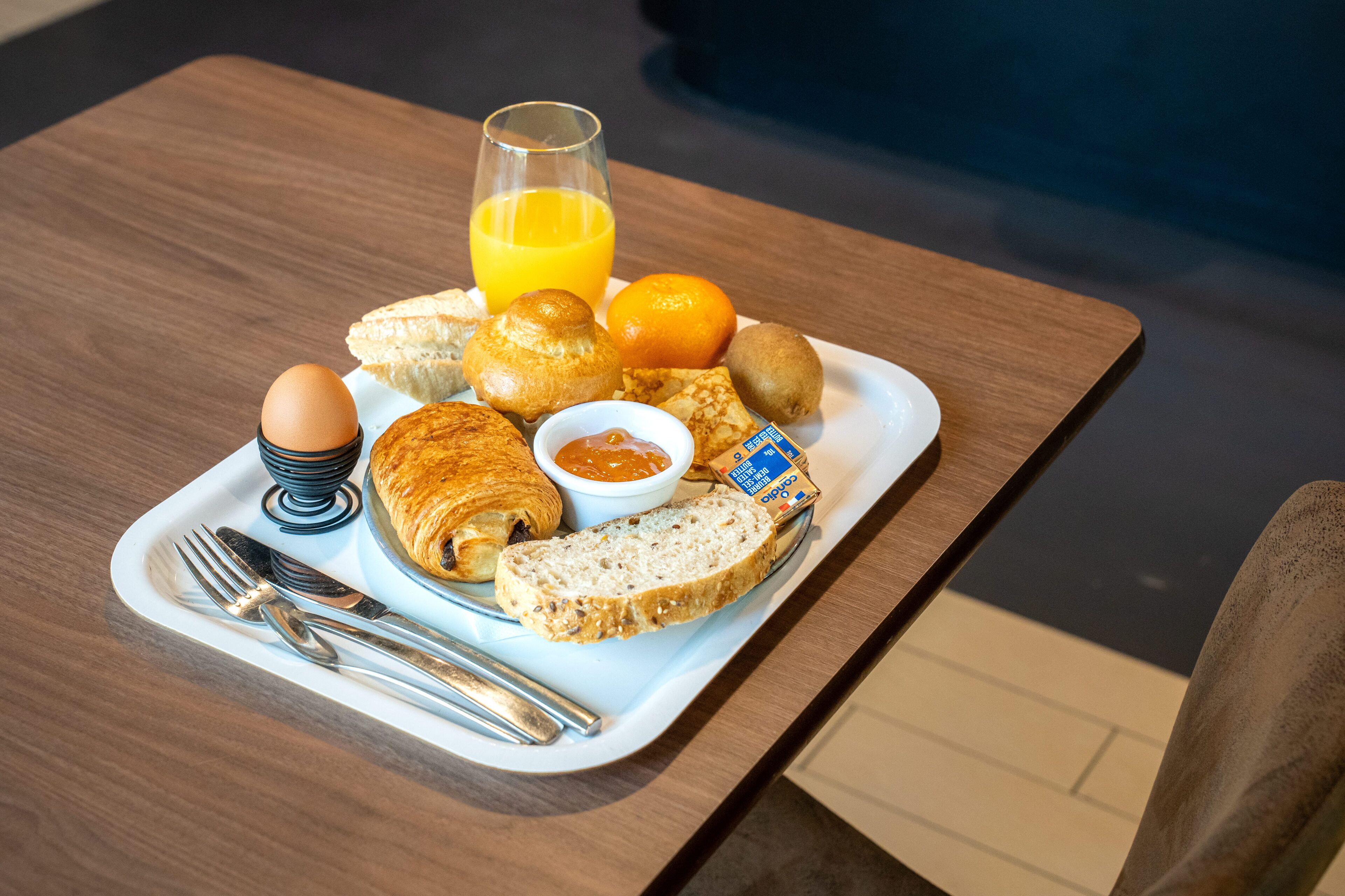 A breakfast tray with croissants, bread, an orange, a kiwi, a glass of orange juice, a boiled egg, and a small bowl of jam.