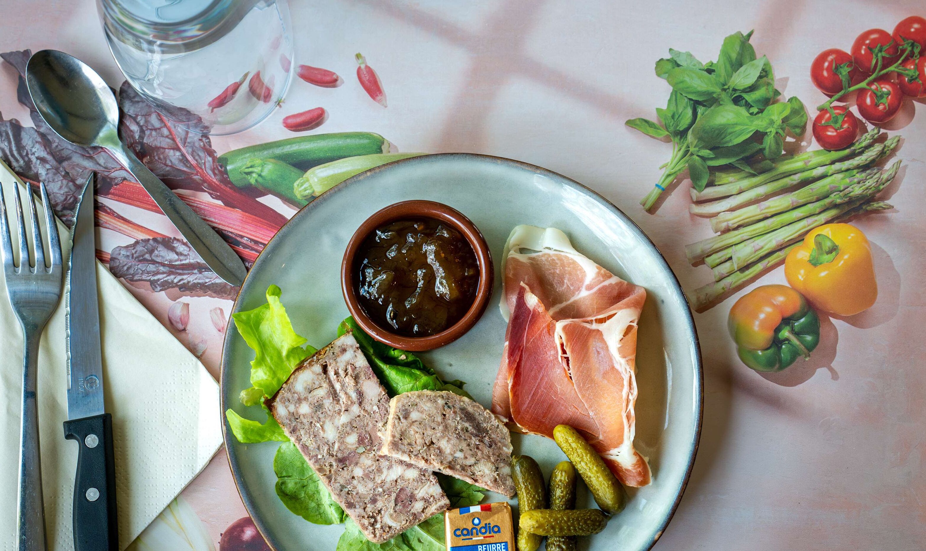 Un plato con diversos alimentos, incluyendo pan, jamón, encurtidos y un bol de salsa, junto con verduras como lechuga y tomates.