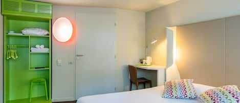 A hotel room with a bed, a desk, a chair, a green wardrobe, and a round wall light.