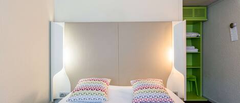 A hotel room with a bed, two patterned pillows, a bedside lamp, a green storage unit, and a white headboard.