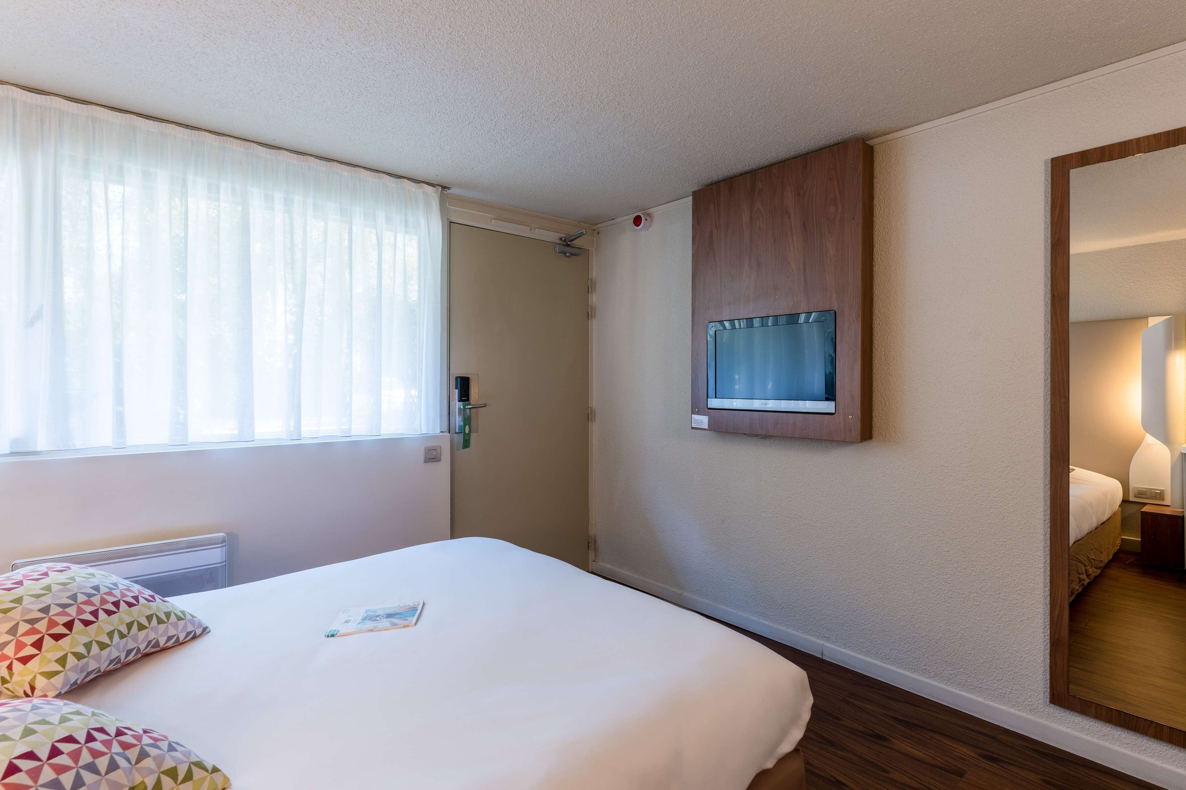 A hotel room with a bed, a TV mounted on the wall, a window with curtains, and a mirror reflecting part of the room.