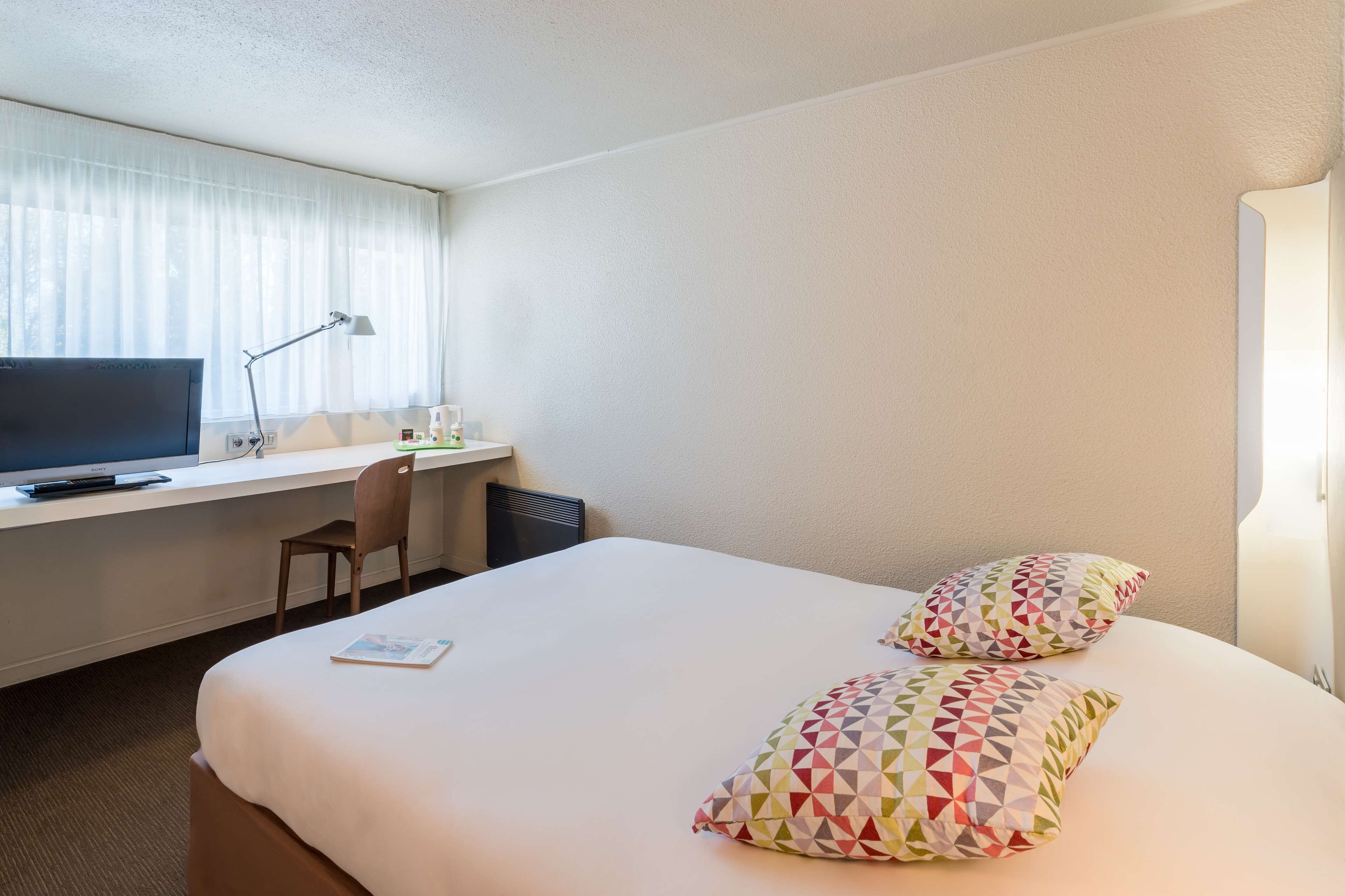 Ein Hotelzimmer mit einem Bett, einem Schreibtisch mit Computer, einem Stuhl und einem Fenster mit Vorhängen.