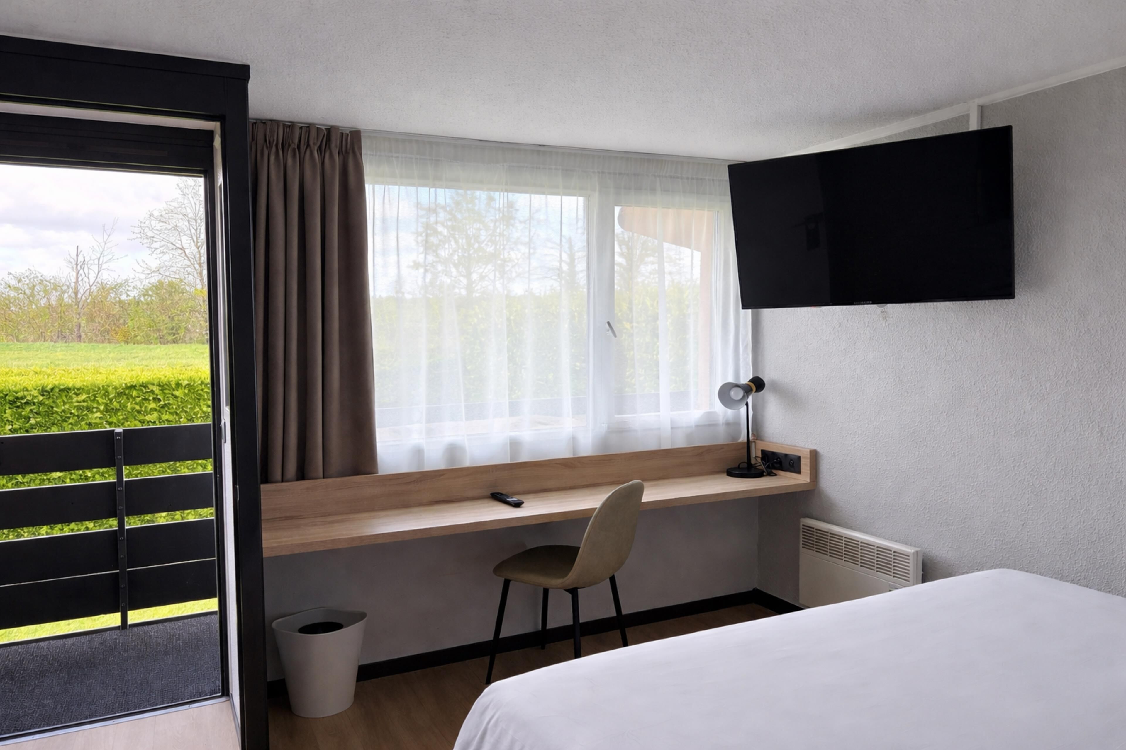 Una habitación de hotel con una cama, un escritorio con silla, un televisor montado en la pared y una ventana con cortinas.