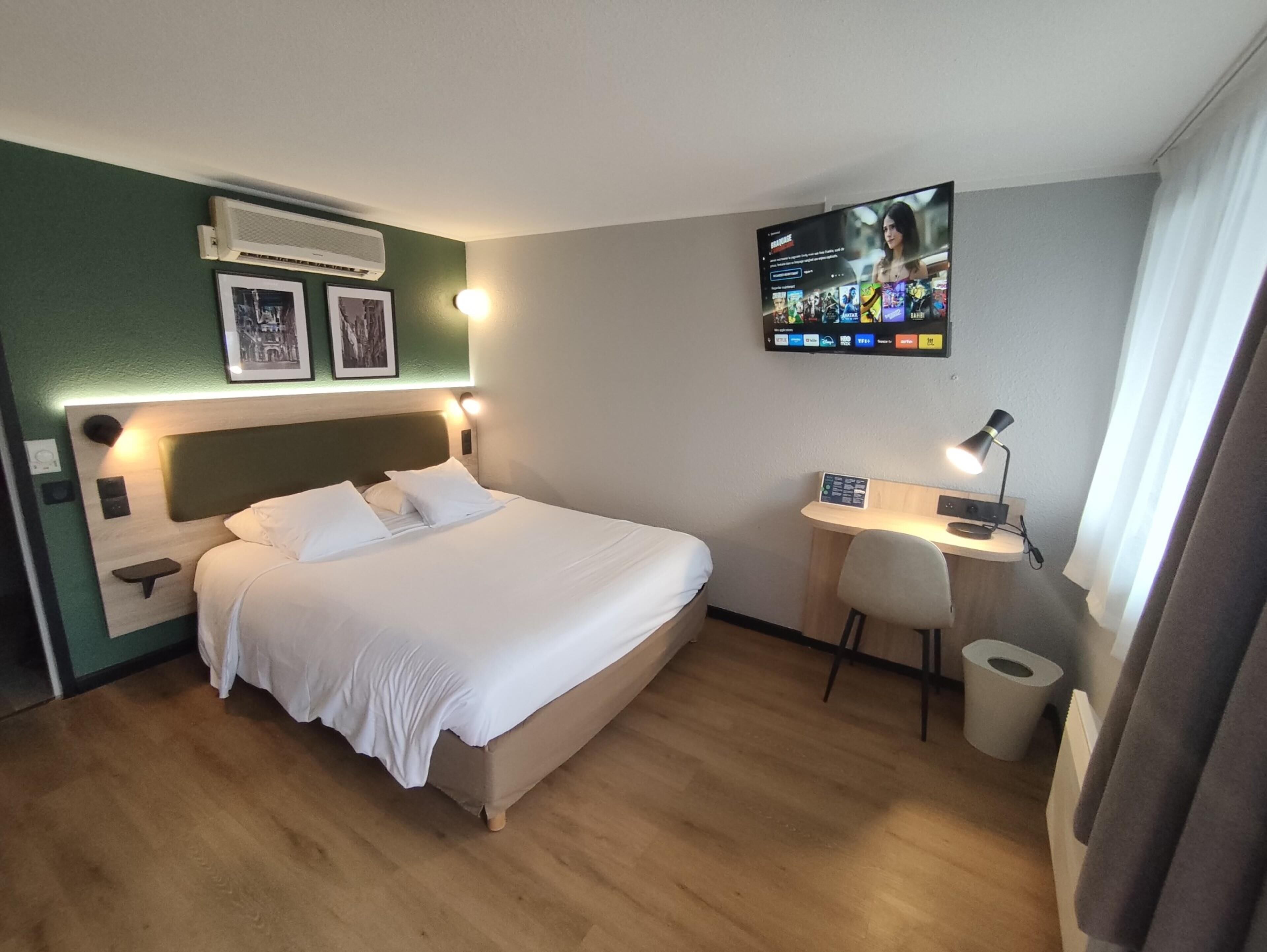 A hotel room with a bed, a desk, a chair, a TV, and a wall-mounted air conditioner.