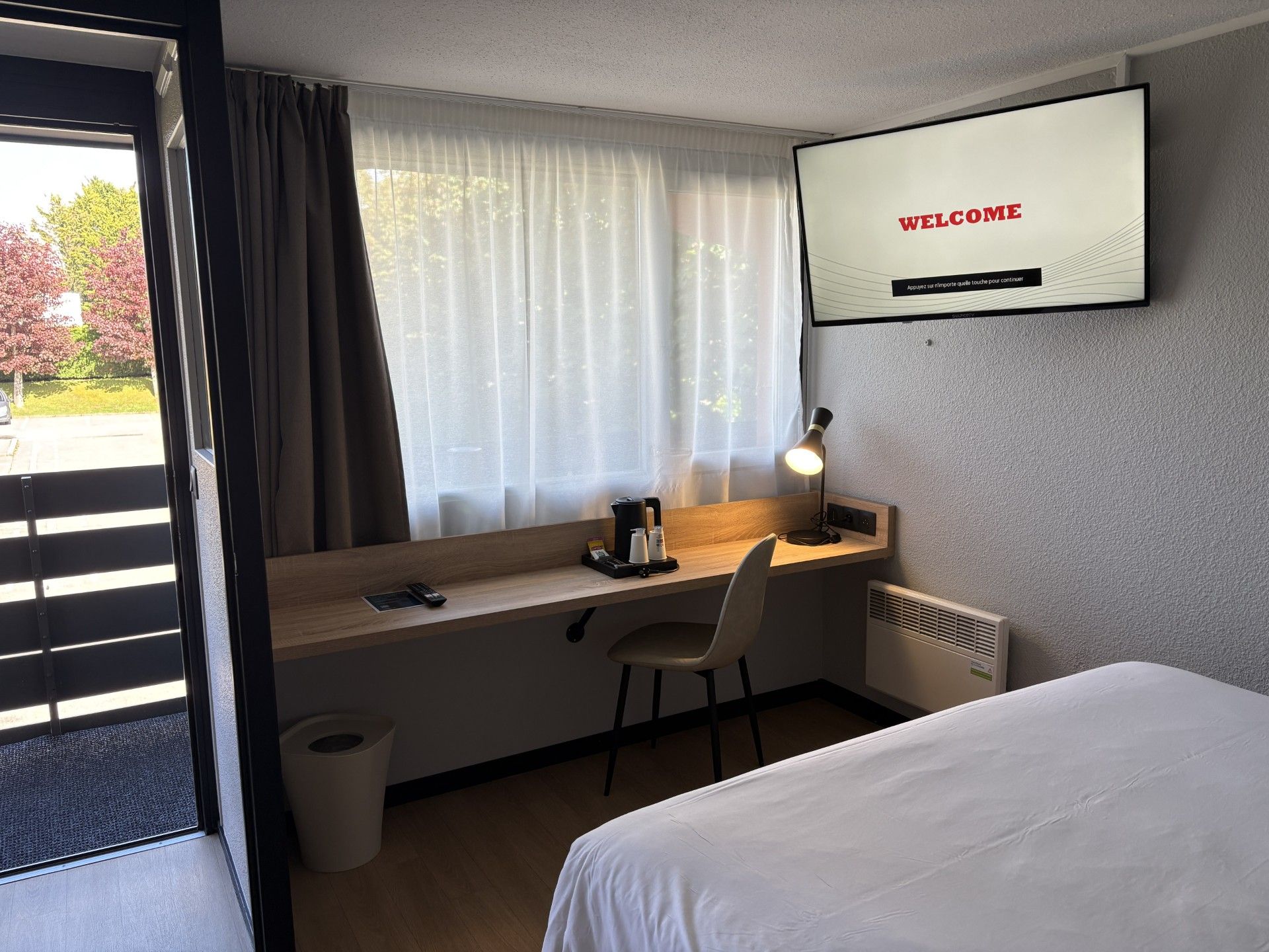 A hotel room with a bed, a desk with a chair, a television displaying 'WELCOME' message, a window with curtains, and a radiator.