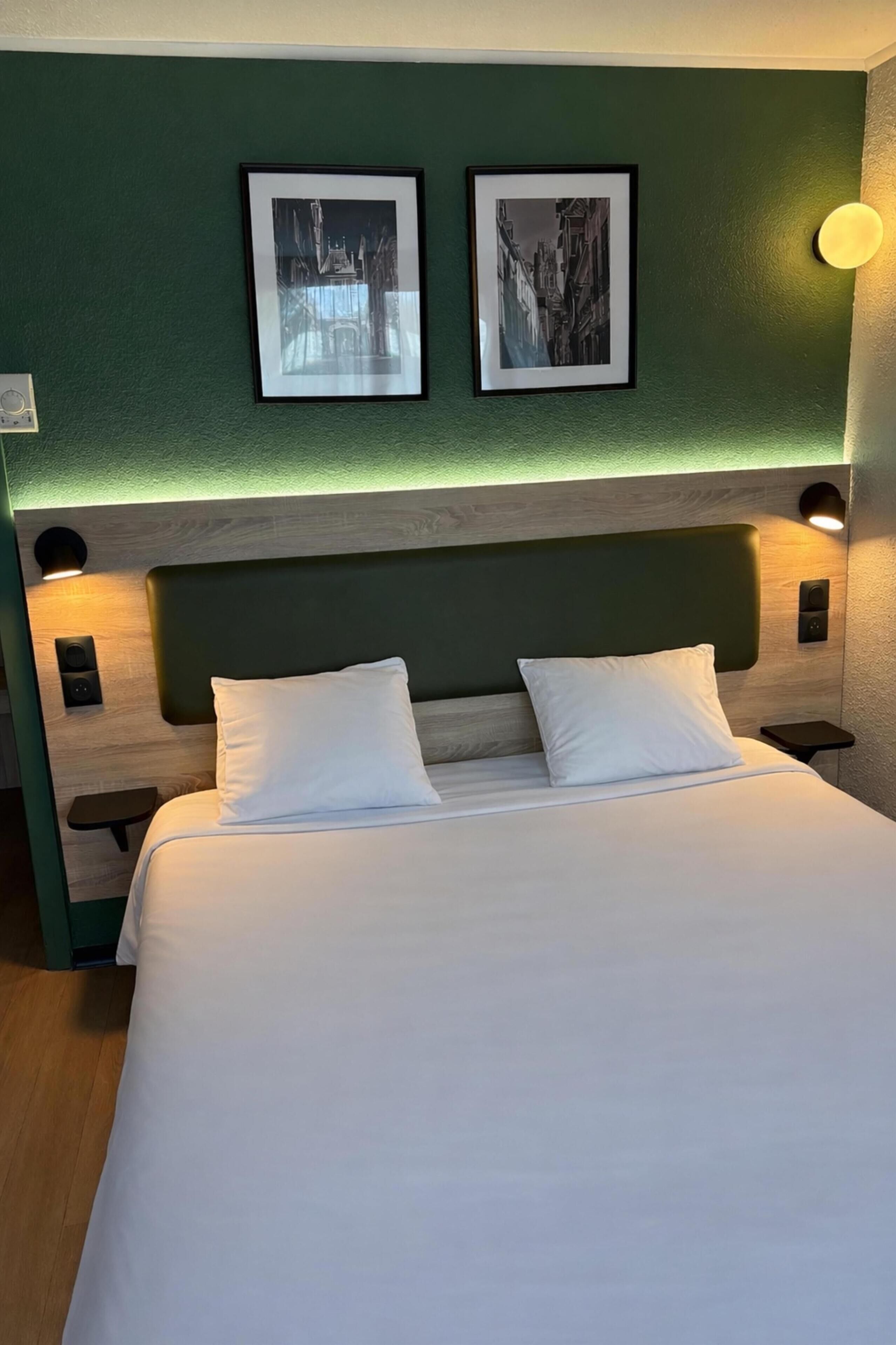 Una cama bien tendida con sábanas blancas, una pared verde y dos cuadros colgados encima.