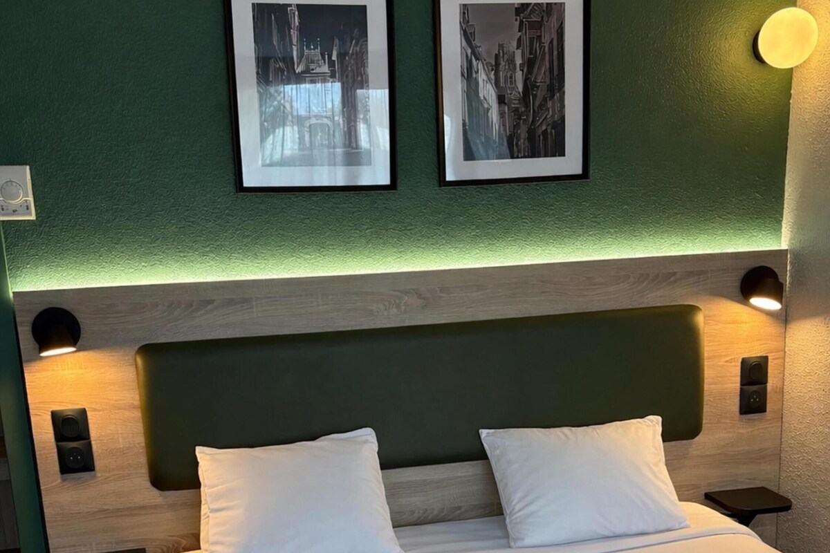 Un lit bien fait, avec du linge de lit blanc, un mur vert et deux tableaux encadrés au-dessus.