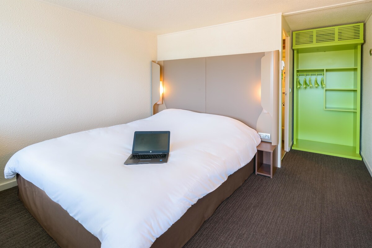 Una habitación de hotel con cama, una laptop sobre la cama, un armario verde y una mesita de noche.