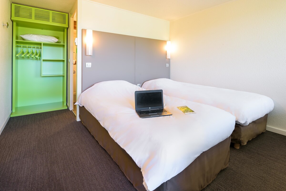 Una habitación de hotel con dos camas, un armario verde y un ordenador portátil sobre una de ellas.