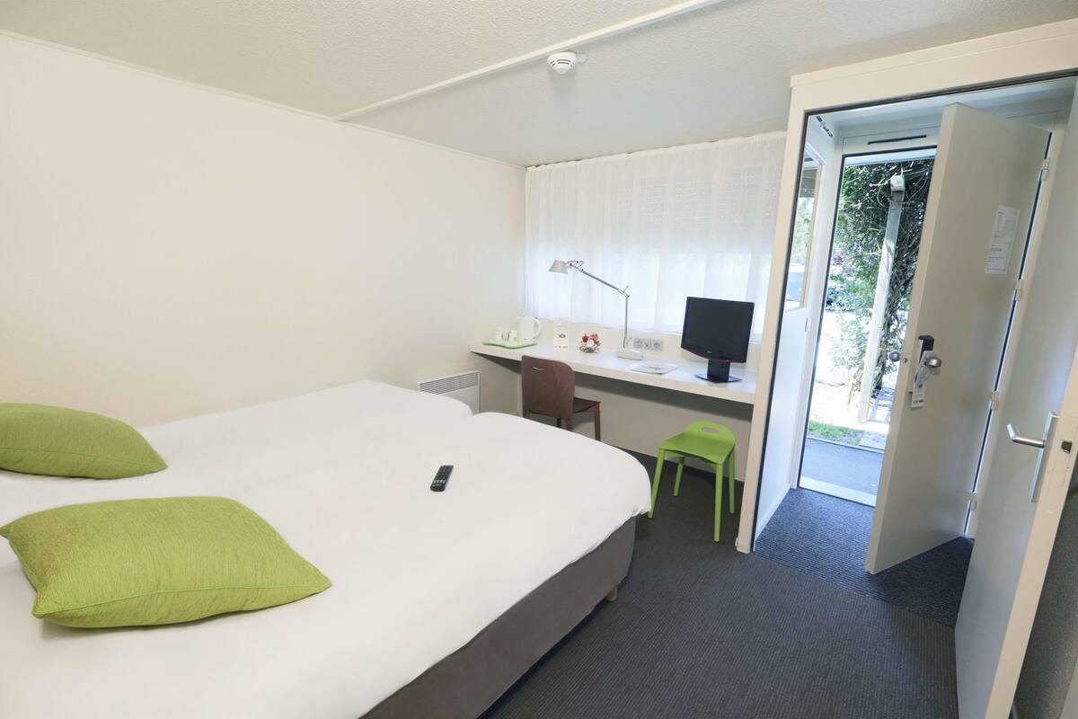 A hotel room with a bed, a desk with a computer, a chair, and a door leading to a balcony.