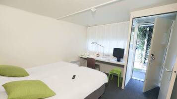 A hotel room with a bed, a desk with a computer, a chair, and a door leading to a balcony.