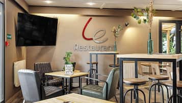 A restaurant interior with a modern design, featuring a flat-screen TV mounted on the wall, wooden tables and chairs, and a decorative plant arrangement.
