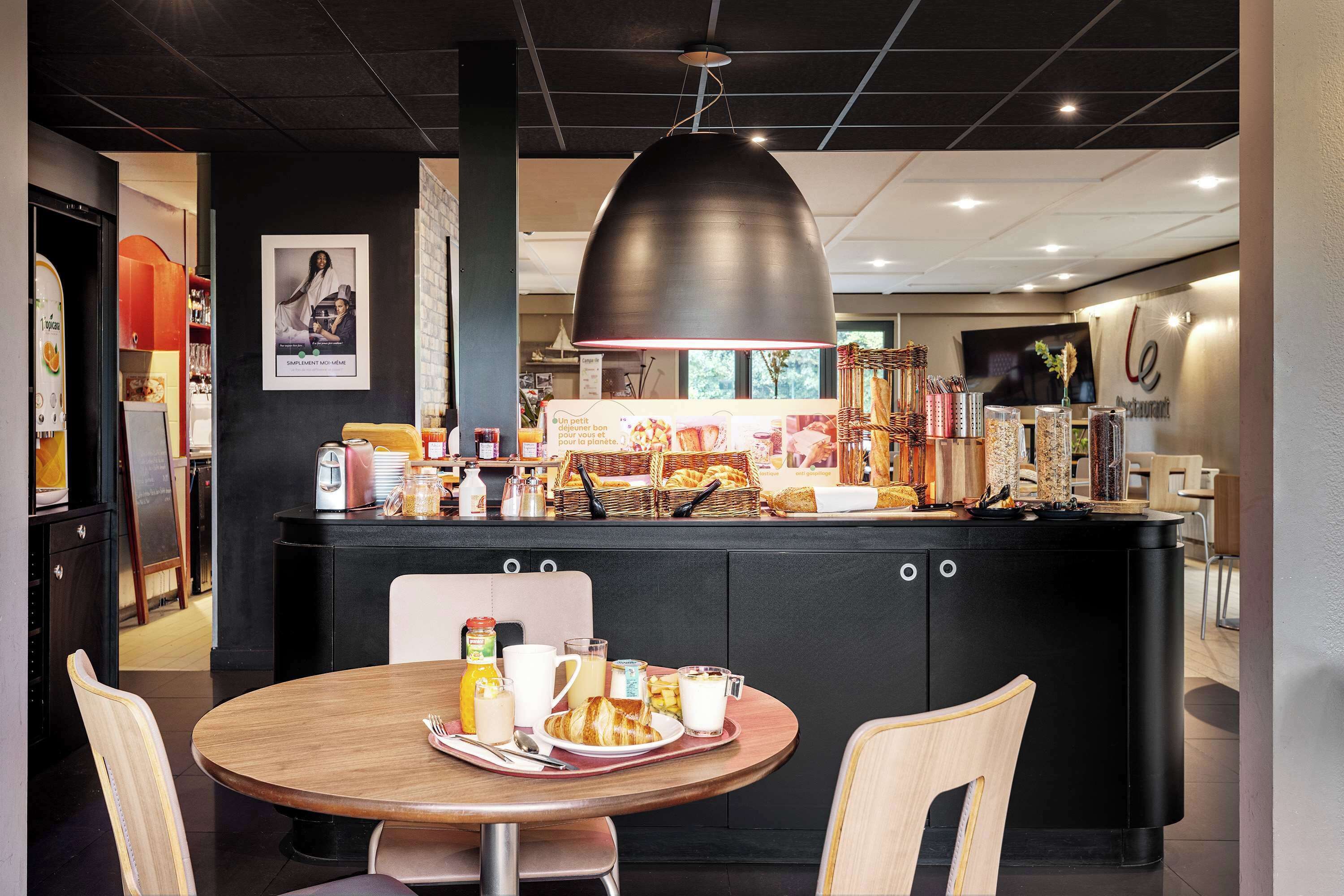 A modern cafe with a central counter, a round wooden table with breakfast items, and a large pendant light.