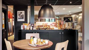 A modern cafe with a central counter, a round wooden table with breakfast items, and a large pendant light.