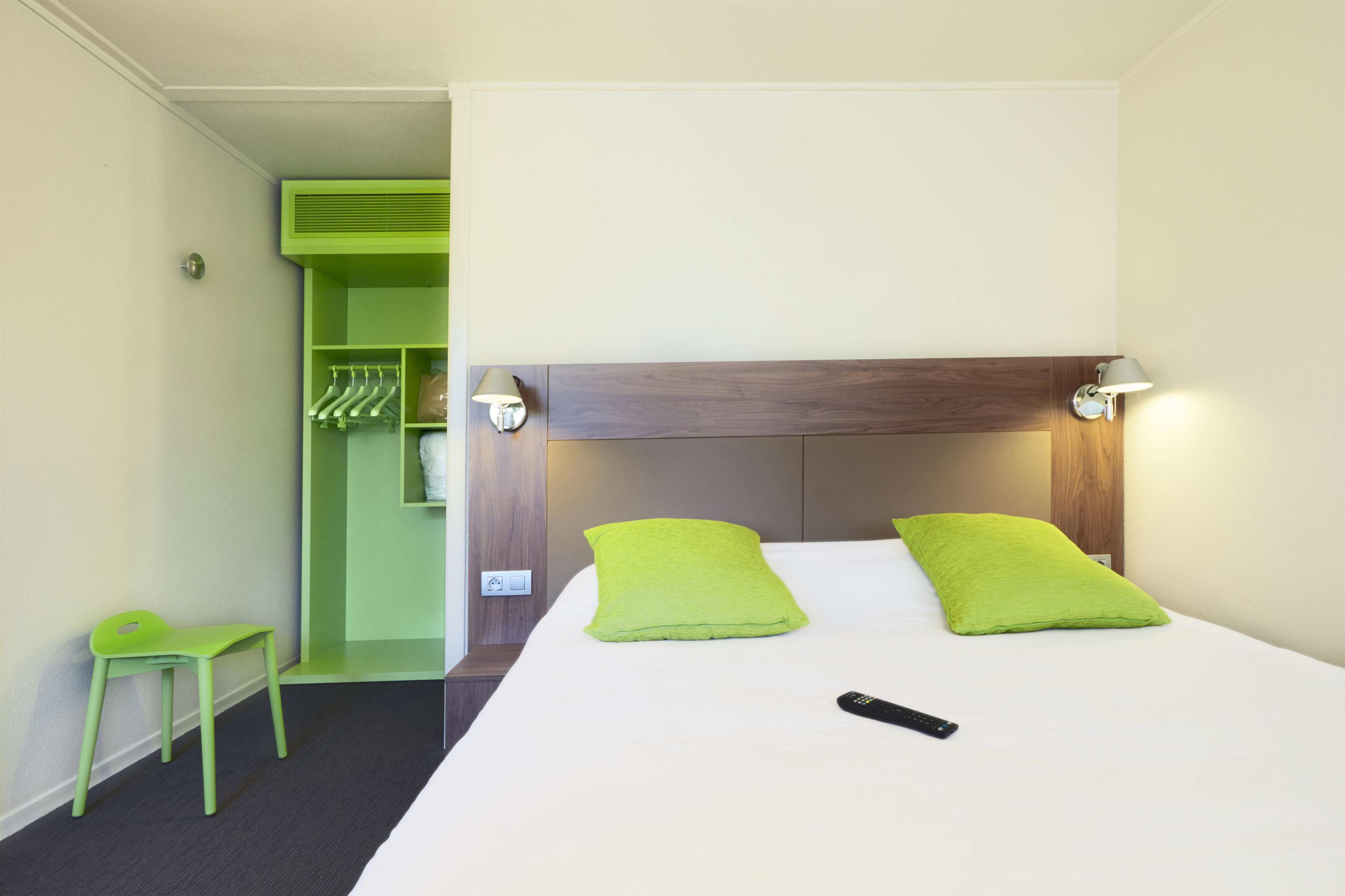 A hotel room with a bed, two green pillows, a remote control, a green chair, and a green wardrobe with hanging clothes.