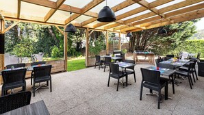 An outdoor patio with wooden beams and black chairs, tables with menus, and a grill area. - Campanile Bethune Fouquieres (Fouquieres-les-Bethune)