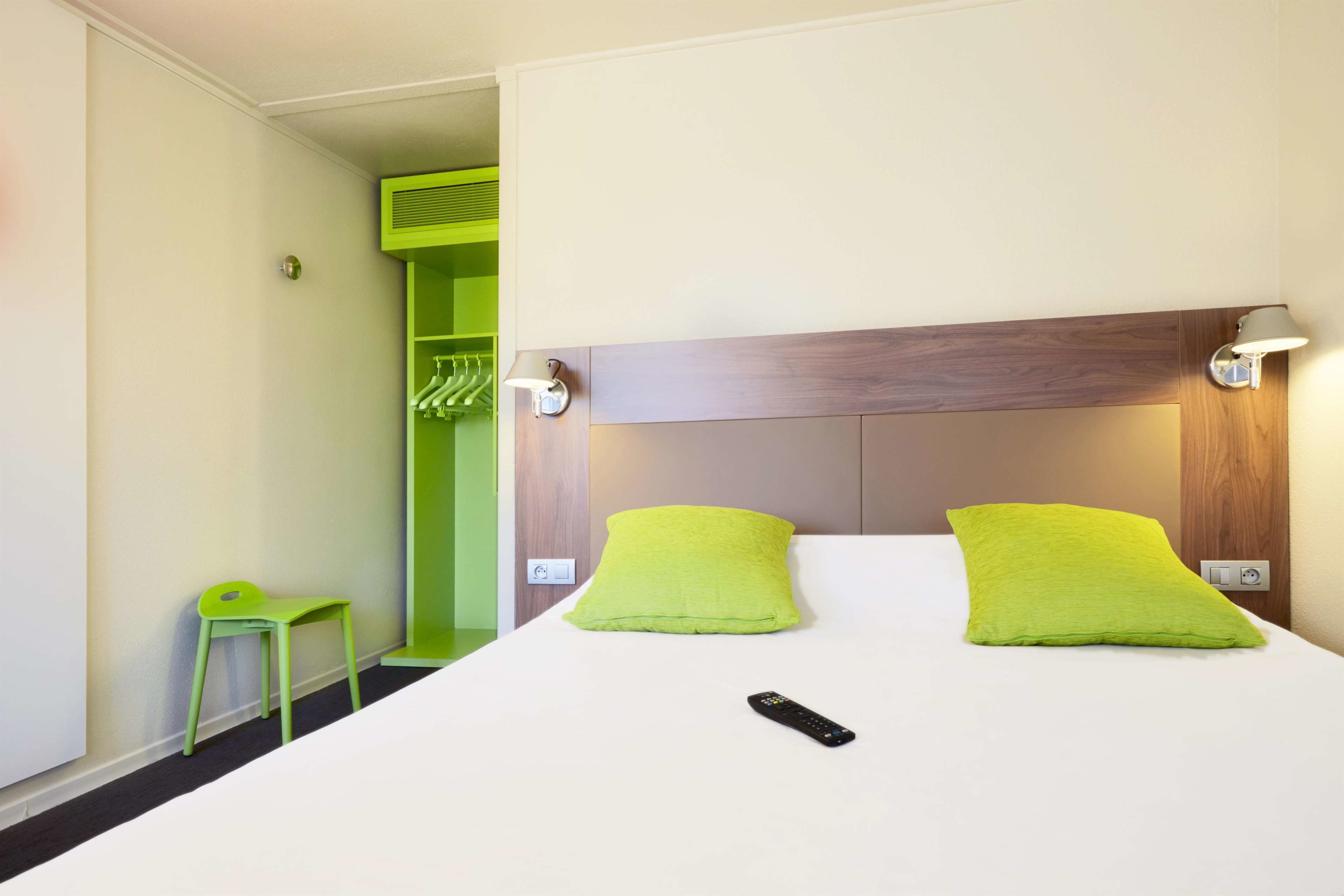 A hotel room with a bed, two green pillows, a green chair, a remote control, and a green closet.