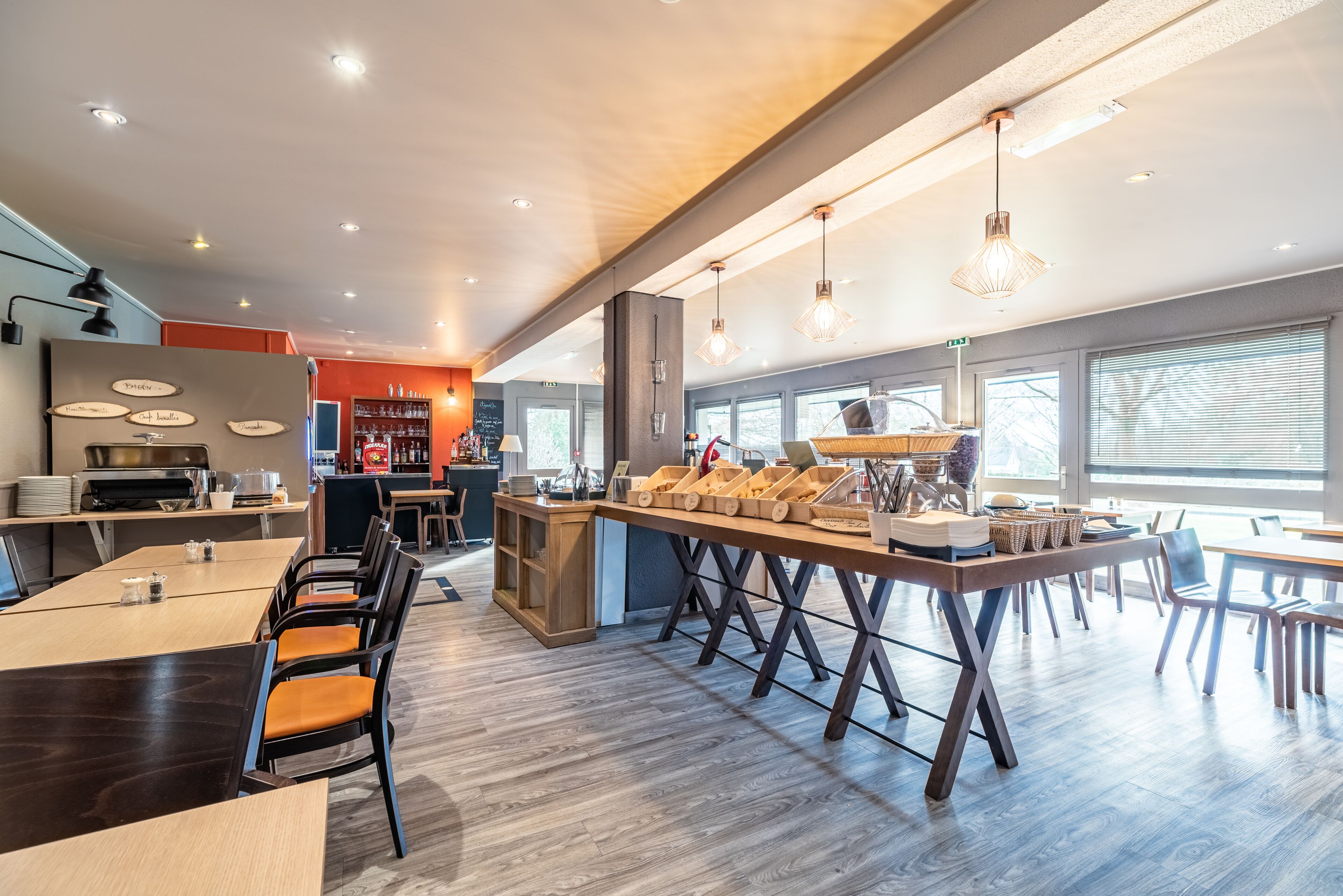 A modern restaurant with wooden tables and chairs, a central bar area, and large windows providing natural light.