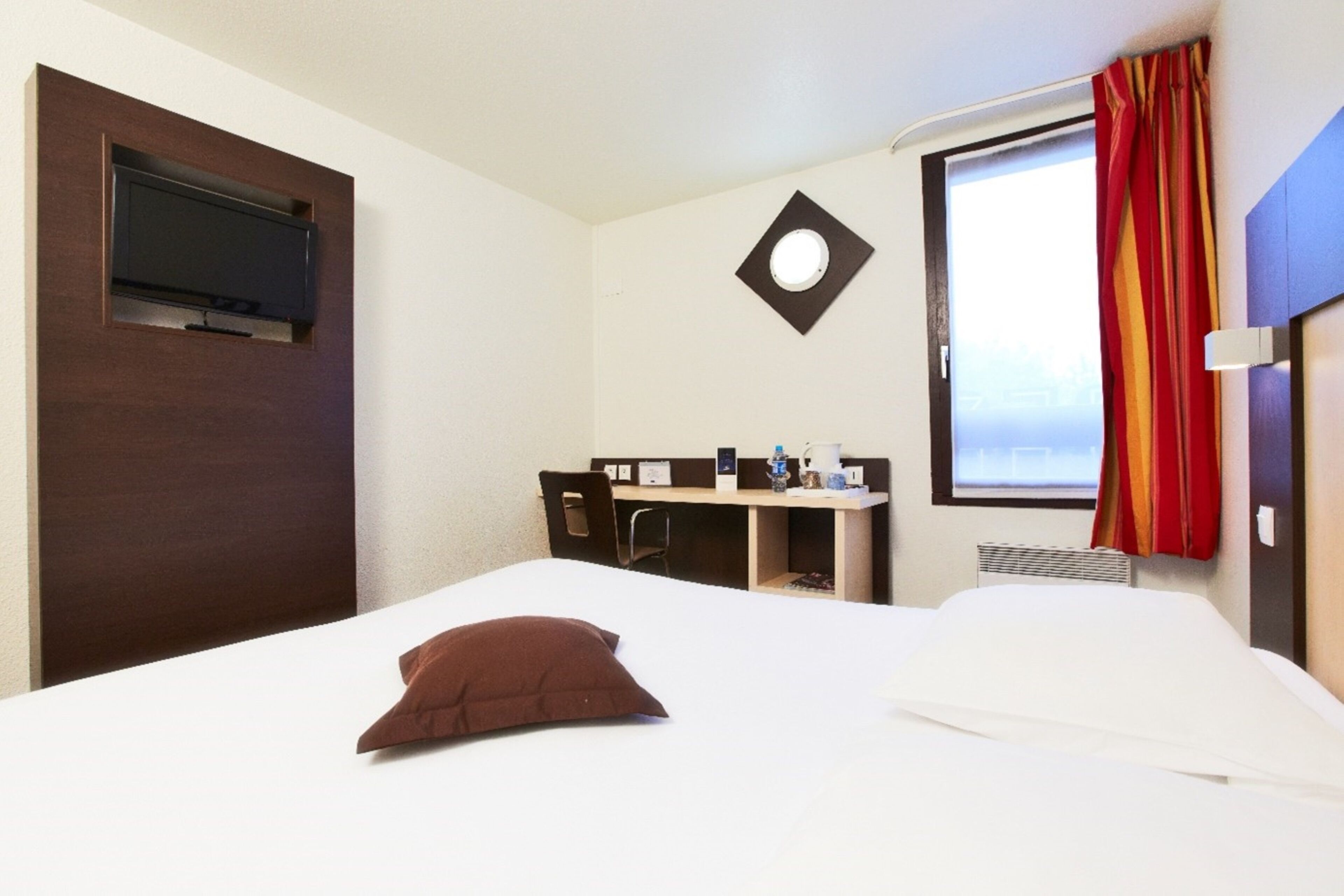 Une chambre d’hôtel comprenant un lit, un bureau avec une chaise, un téléviseur fixé au mur et une fenêtre avec des rideaux.