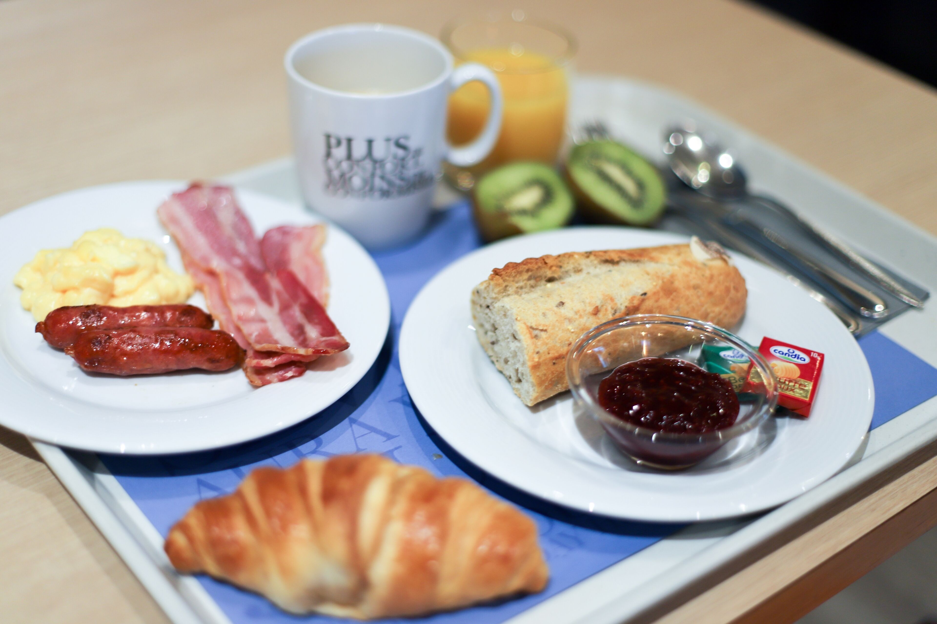 Una bandeja de desayuno con huevos revueltos, tocino, salchicha, tostadas, mermelada, un croissant, una taza de café y un vaso de jugo de naranja.