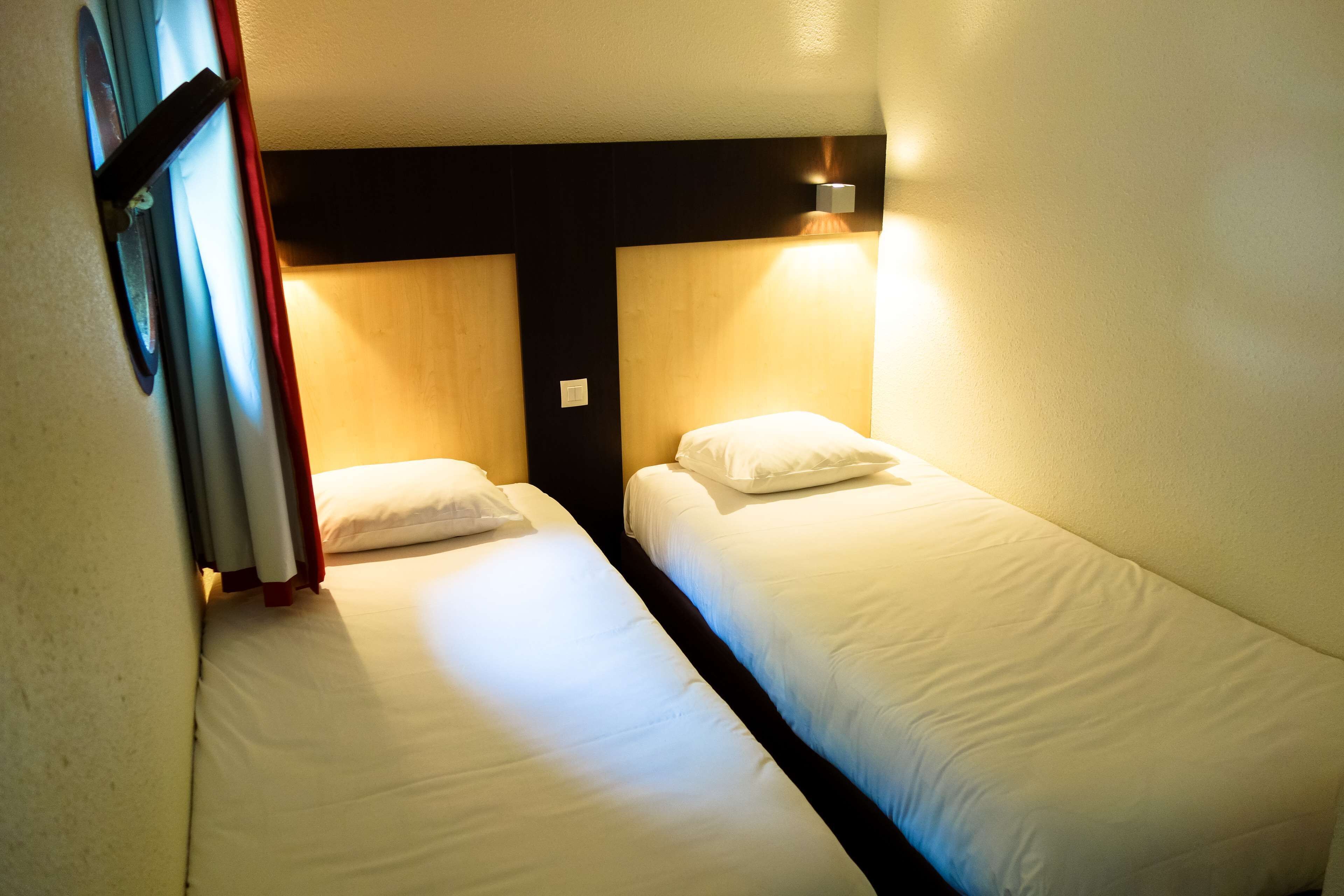 Twee enkele bedden met witte lakens, een houten hoofdbord en een wandlamp.