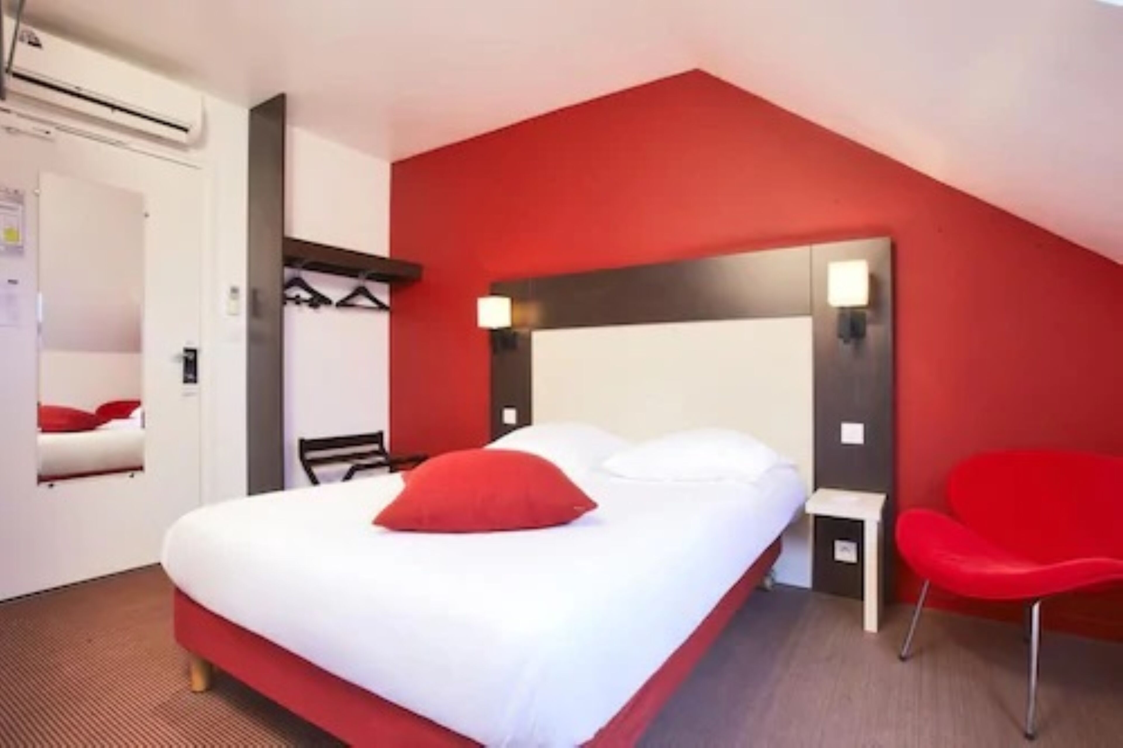 Quarto de hotel com uma cama, uma cadeira vermelha, um porta-roupas e um aplique na parede.