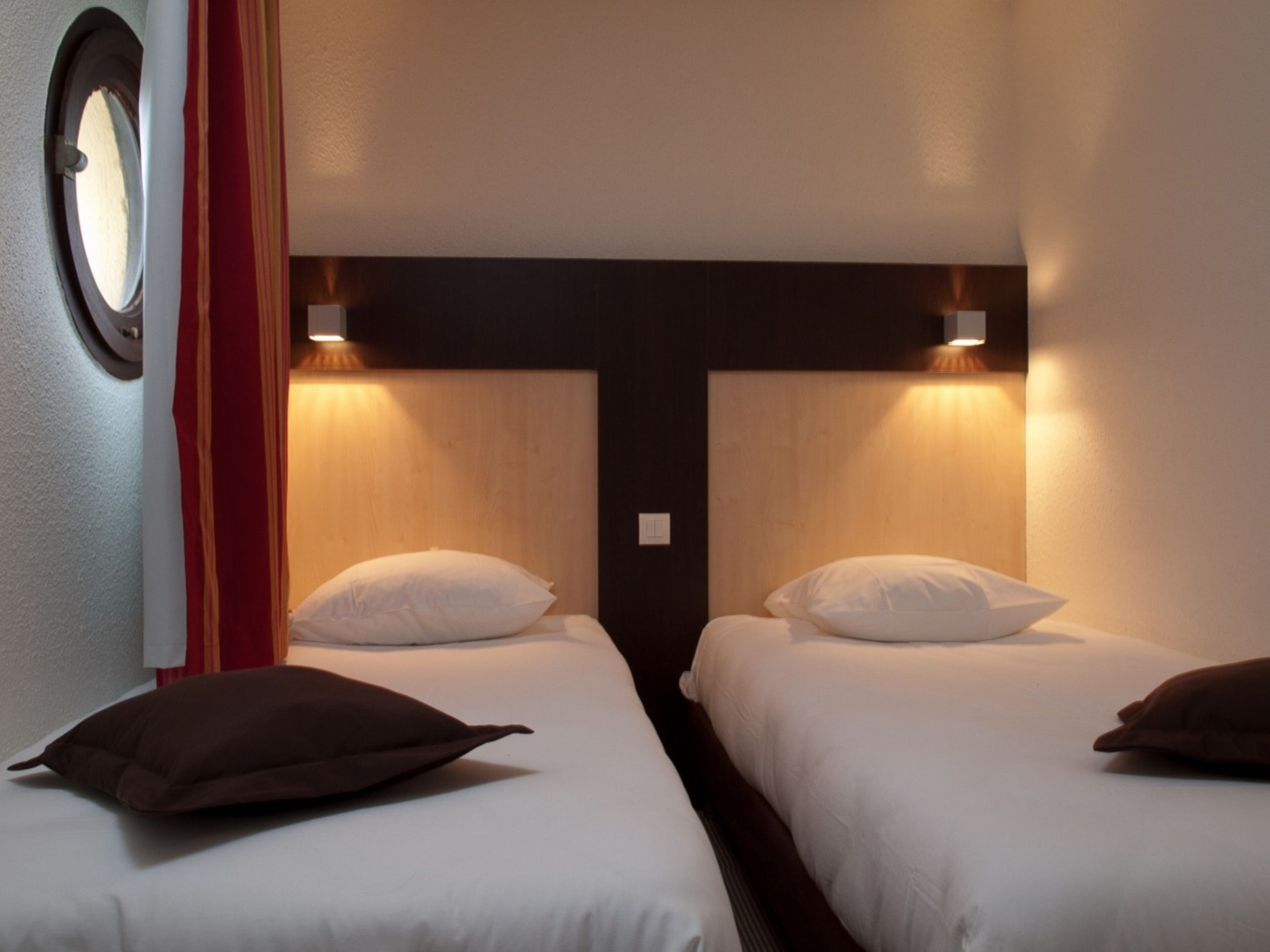 A hotel room with two single beds, each with a pillow and a headboard. There is a window on the left side of the room.