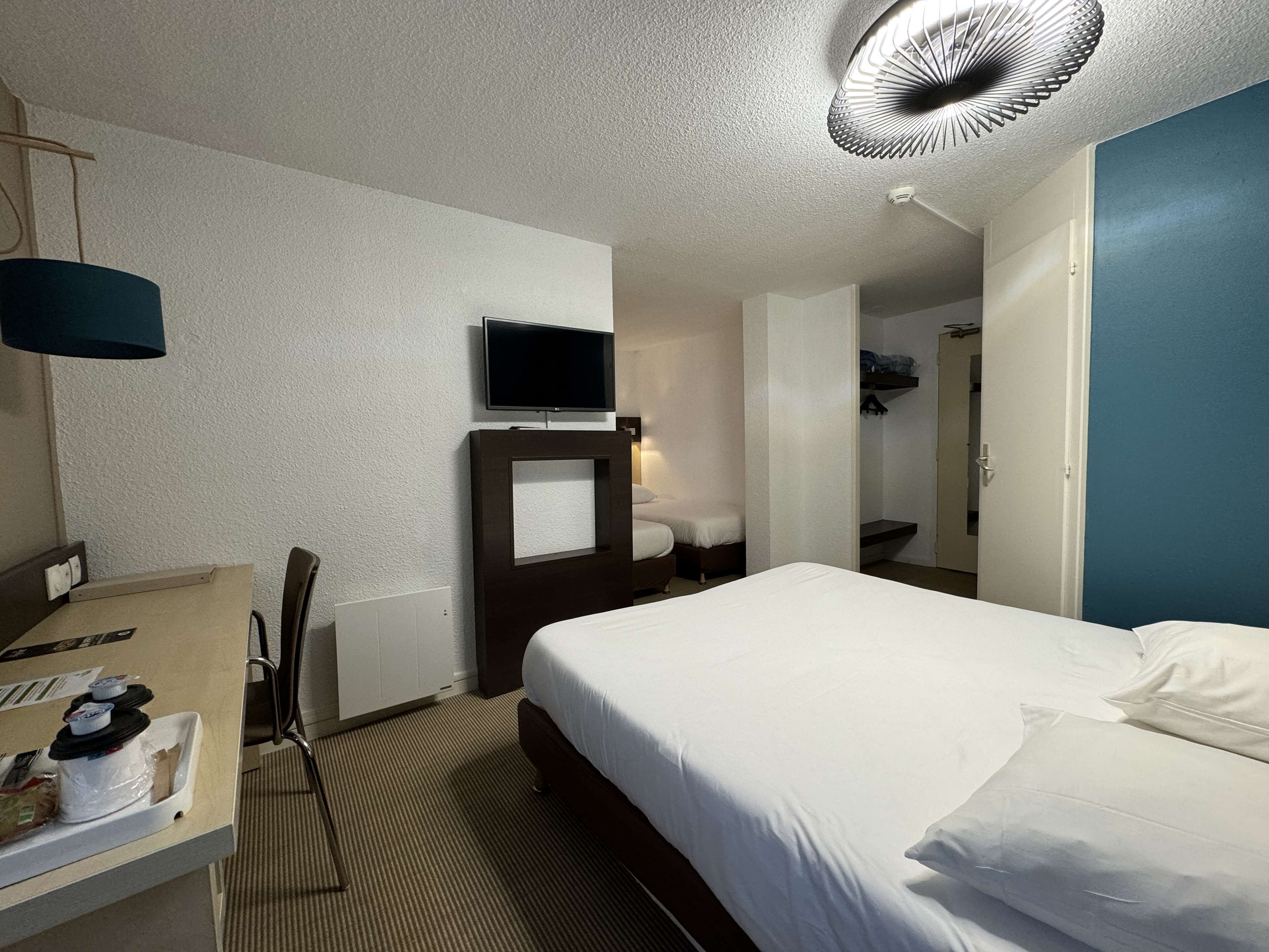 Habitación de hotel con cama, escritorio con silla, televisión montada en la pared y ventilador de techo.