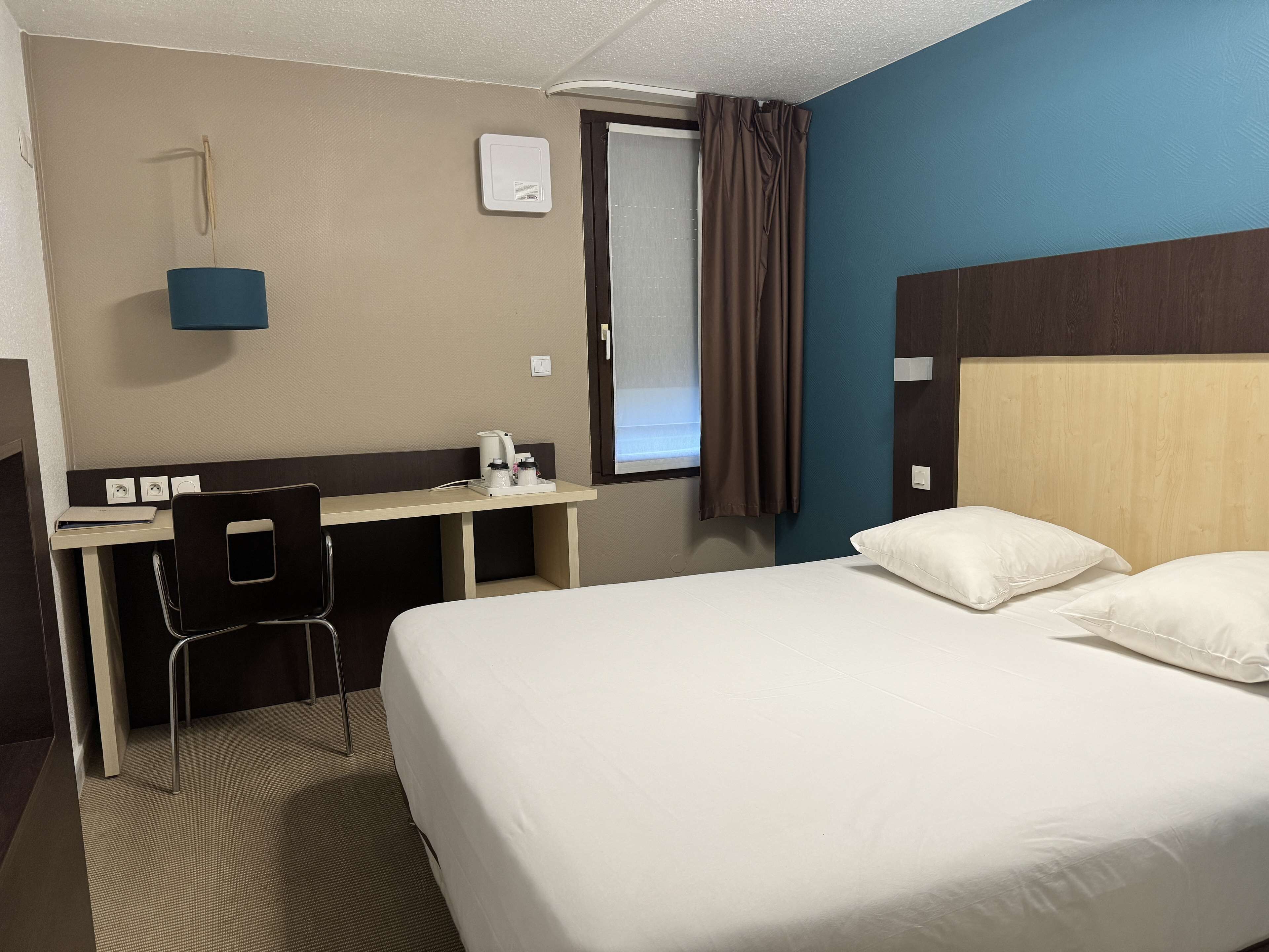 A hotel room with a bed, a desk with a chair, a window with curtains, and a wall-mounted heater.