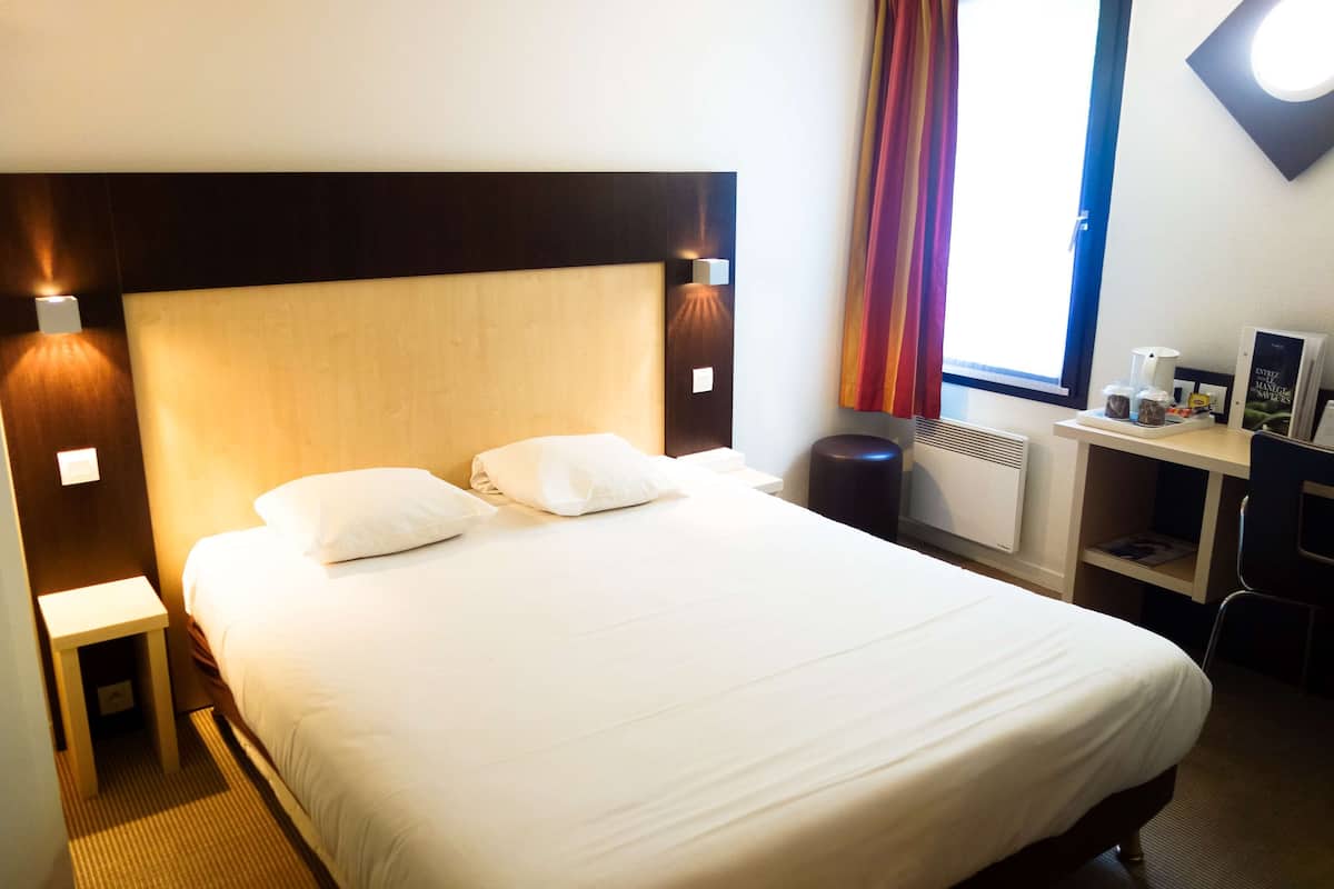 A hotel room with a large bed, bedside tables, a desk with a chair, and a window with curtains.
