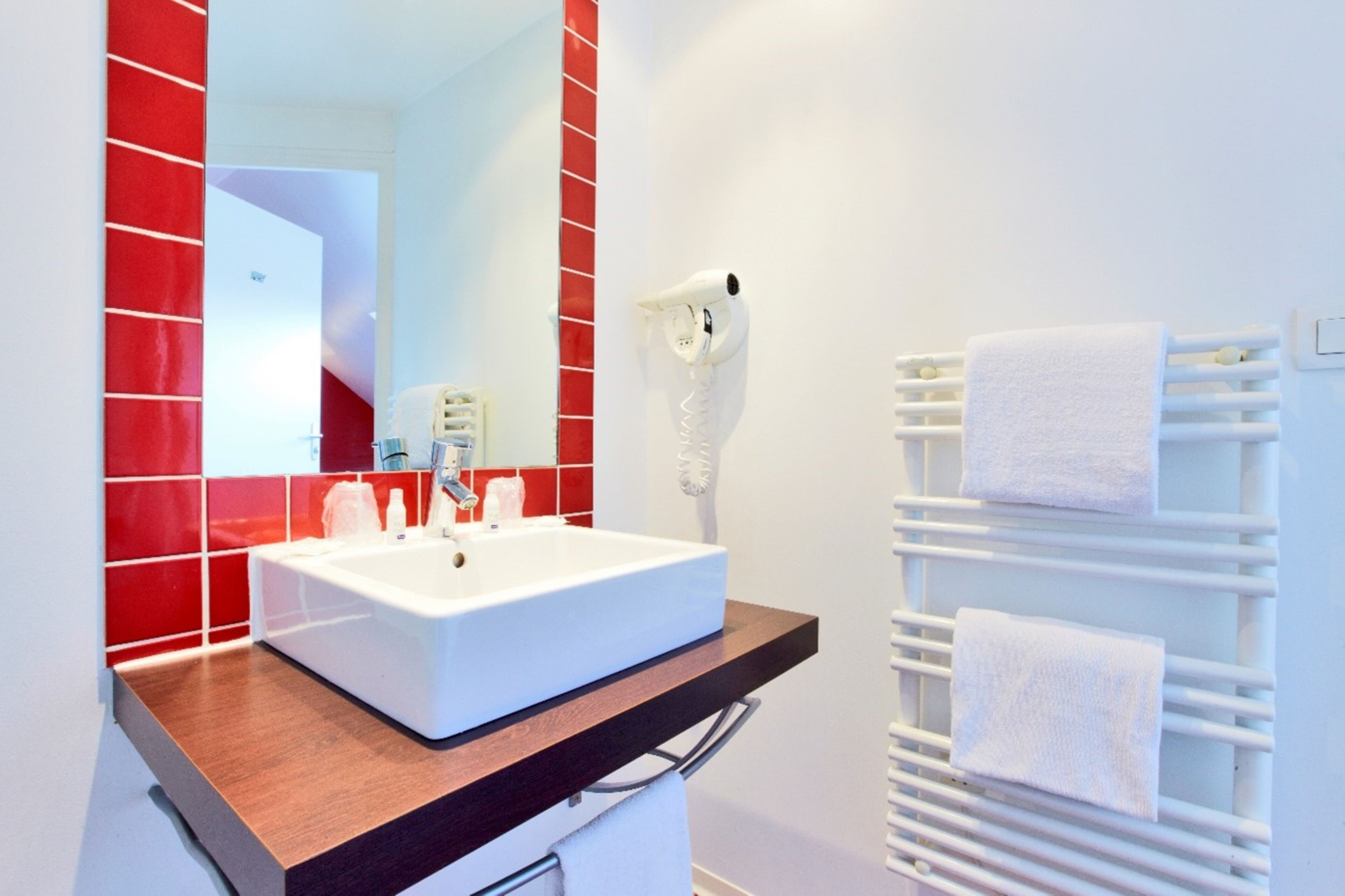 Un baño con un lavabo blanco, paredes de baldosas rojas, un toallero blanco y un secador de pelo montado en la pared.
