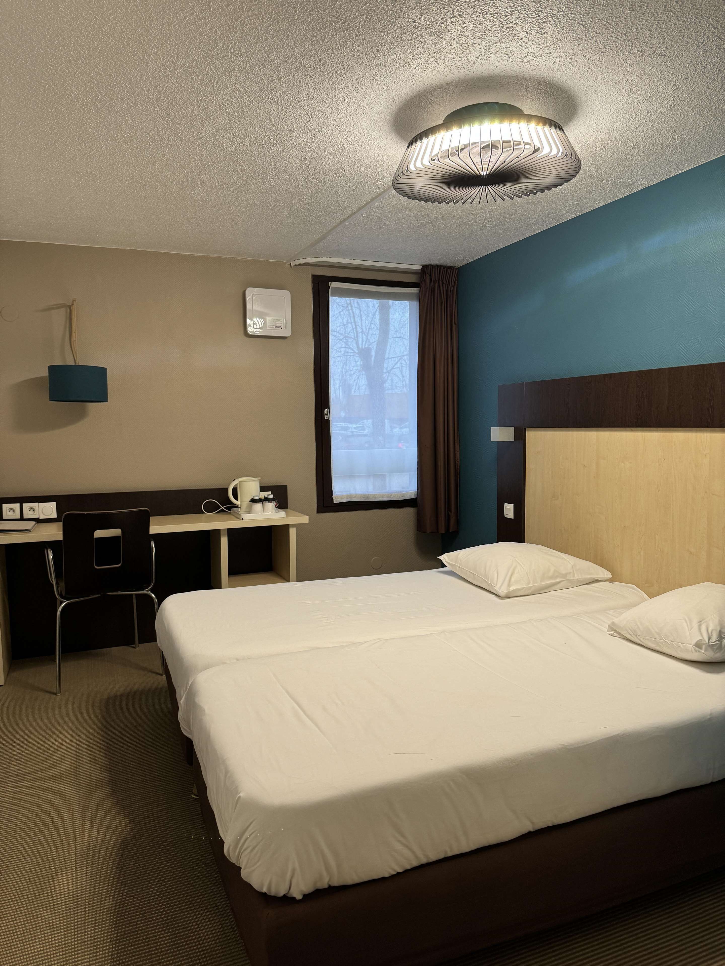 A hotel room with a bed, a desk, a chair, a window with curtains, and a ceiling light fixture.