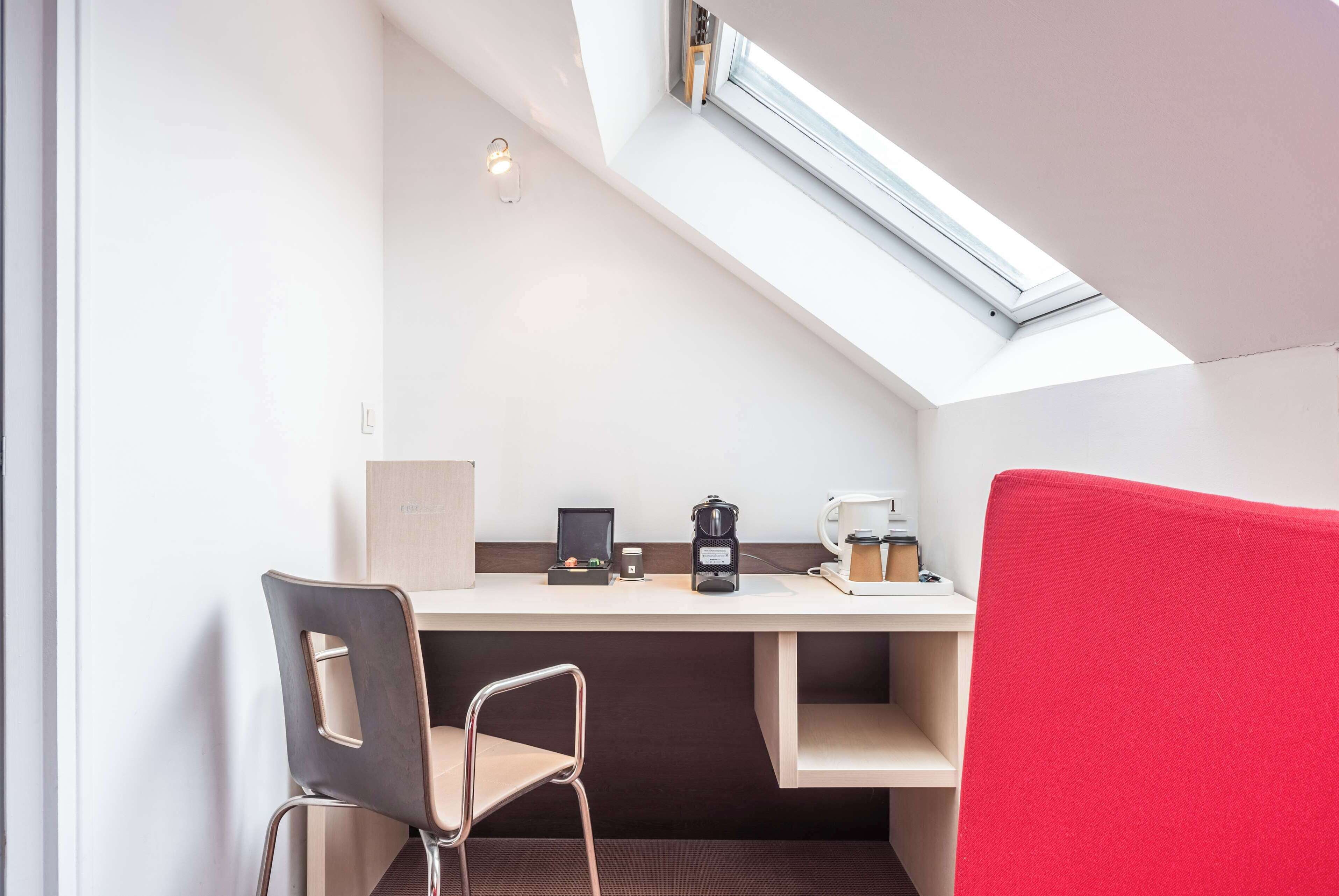 Una habitación moderna con techo inclinado, tragaluz, escritorio con computadora, cafetera y una silla roja.