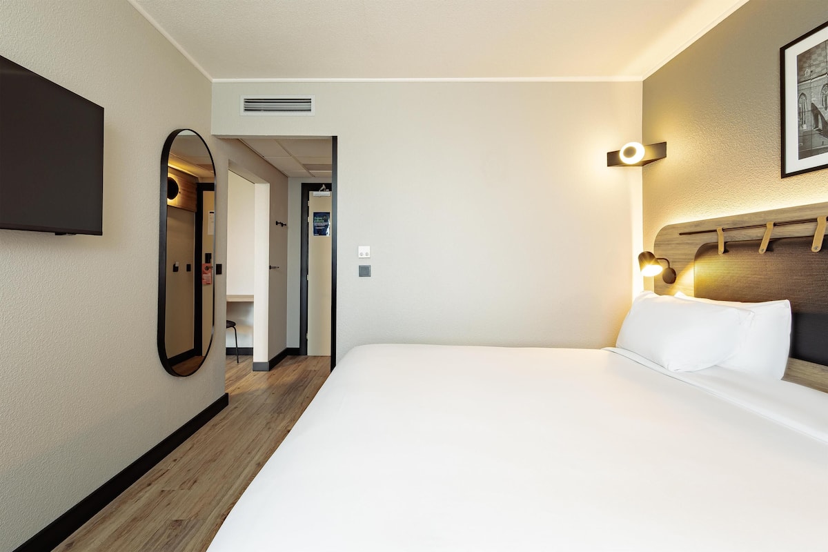 Habitación de hotel con cama, televisor montado en la pared, espejo y cabecero con luces.